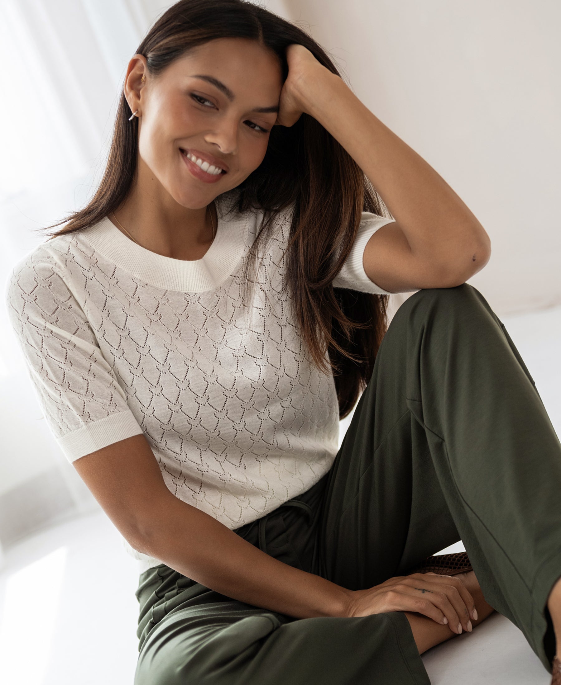 Woman wearing ivory sweater with heart pattern, arm on knee. Sweater by PLEIN PUBLIQUE, style LE BATTANT.
