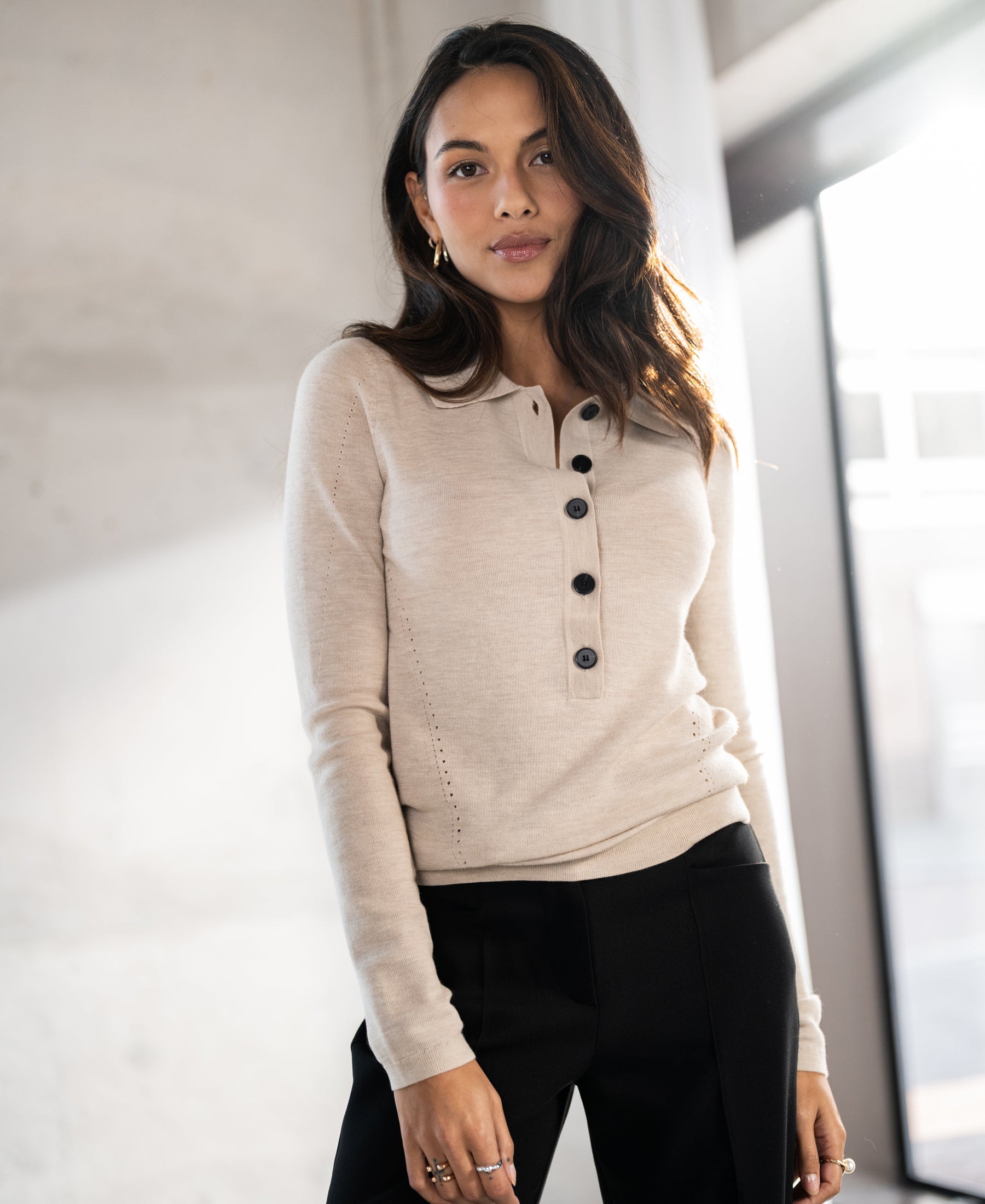 Woman wearing a beige merino wool polo shirt stands by a window, facing forward.