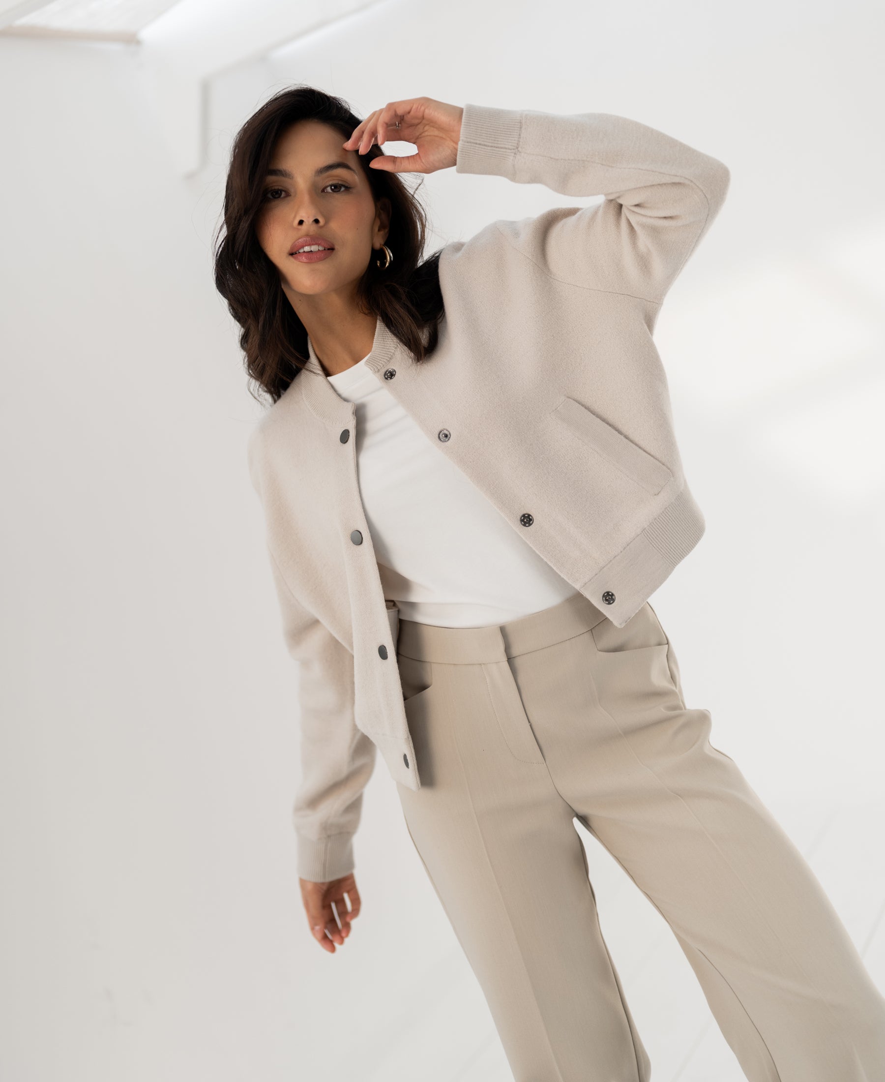 Woman wearing beige LA BOMBA bomber jacket poses indoors with one arm raised, on a white background.