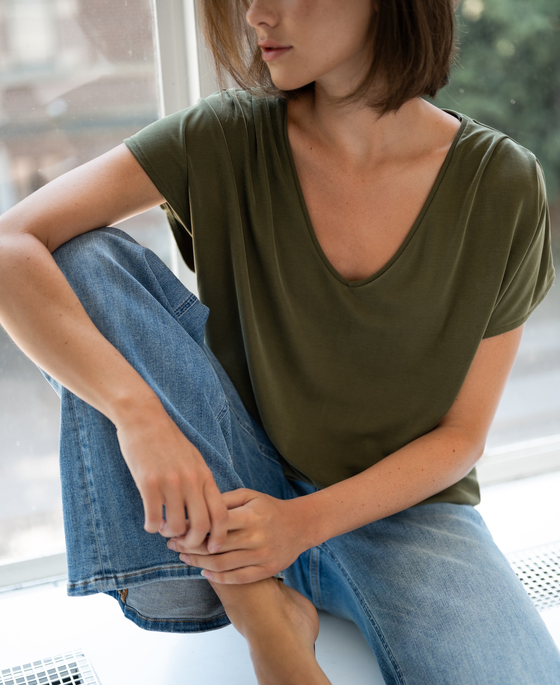 Woman wearing a ribbed olive green LA BONNE T-shirt by PLEIN PUBLIQUE, sitting by a window.