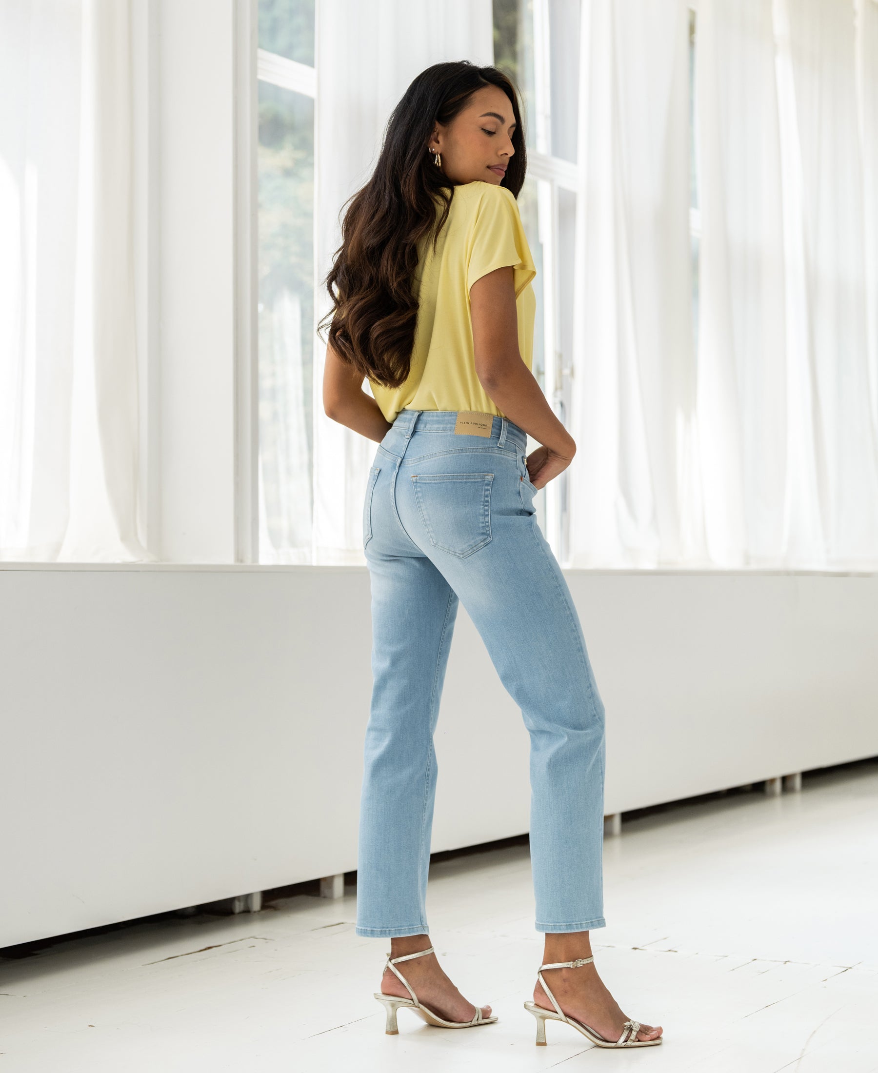 Woman wearing bleached cropped jeans stands indoors sideways with hands in back pockets.