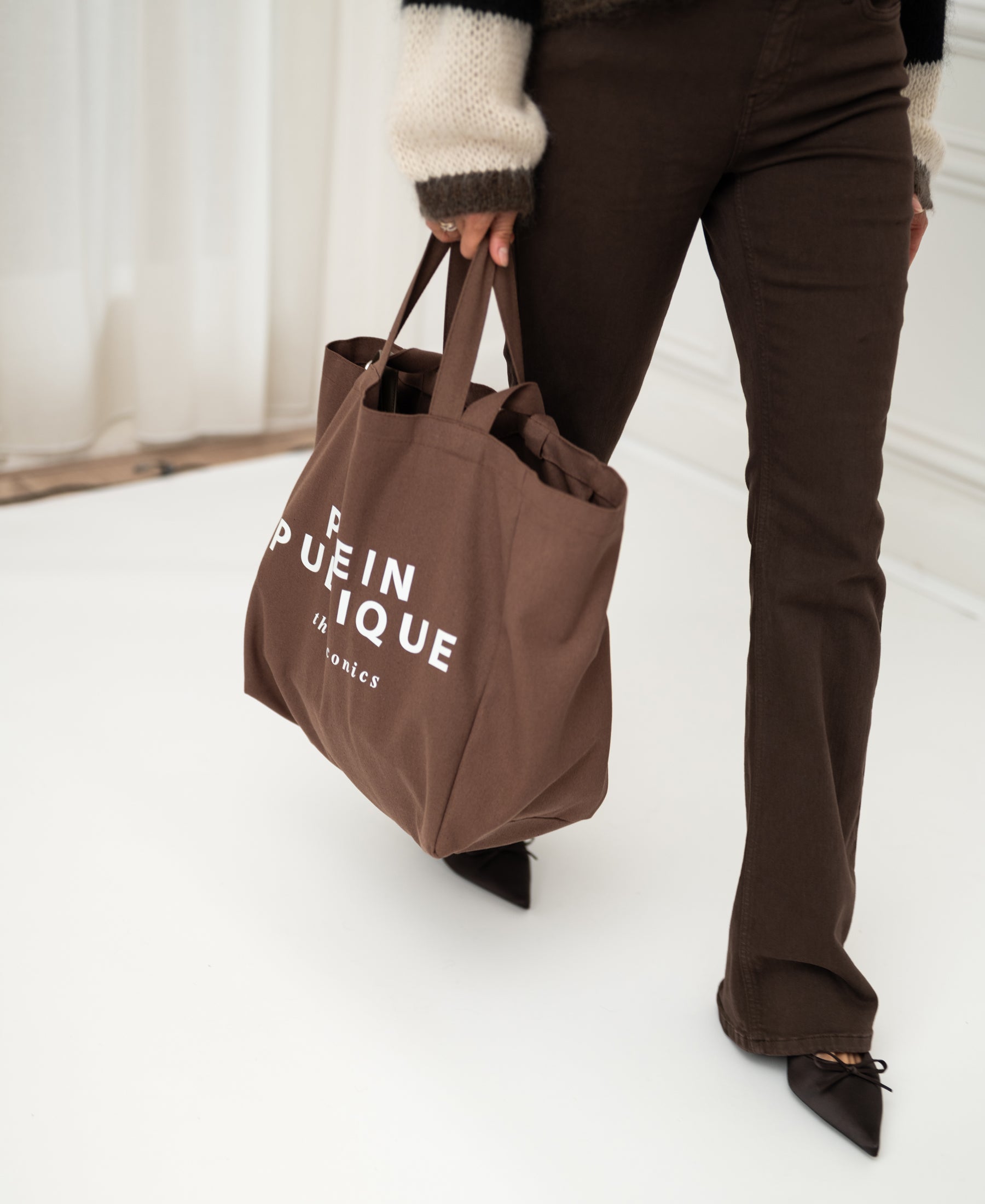 Brown canvas tote bag with "Plein Publique" branding, called "The Bag," shown in a person's hand.