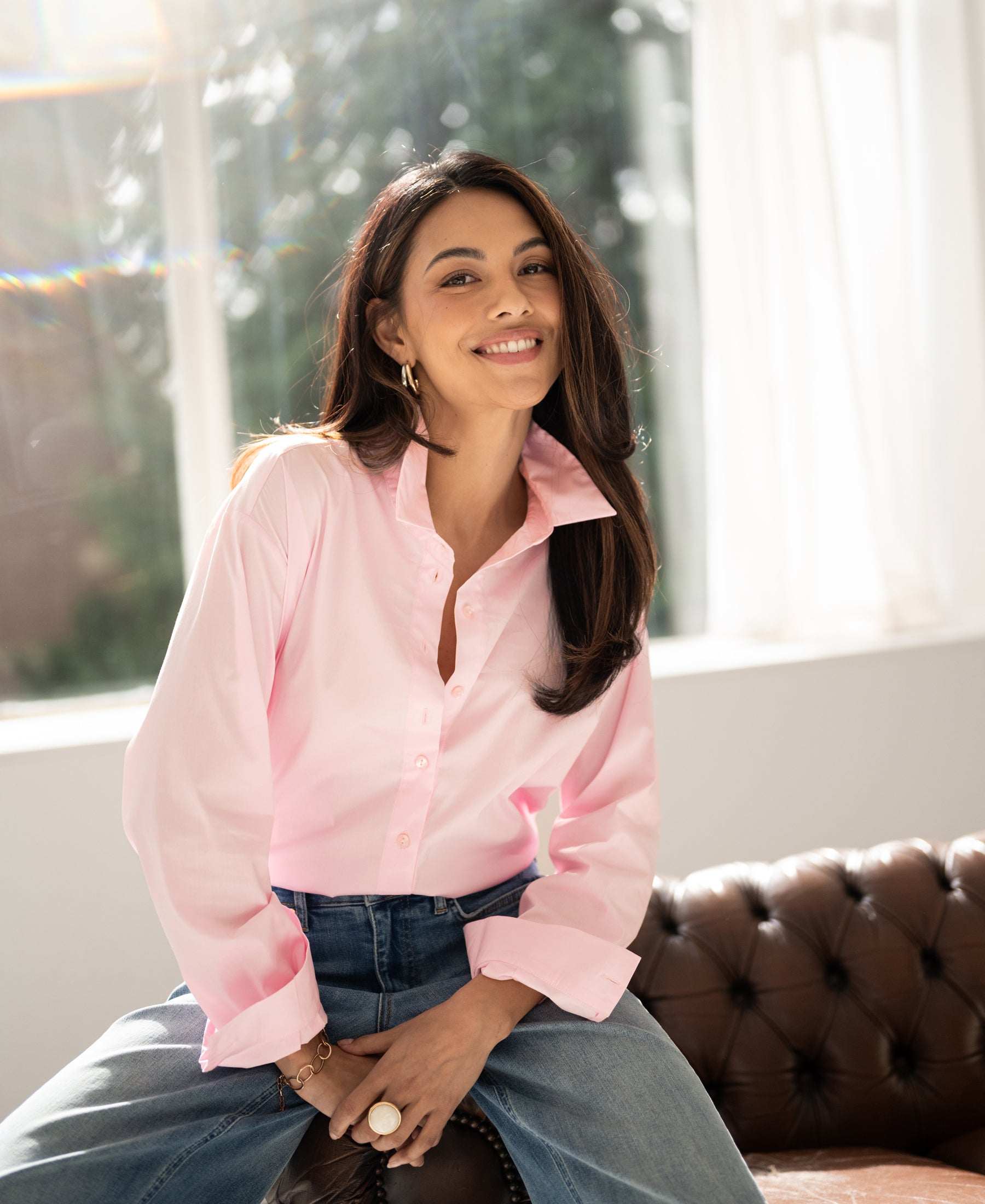 Oversized easy-iron blouse in light pink displayed on a model, shown seated on a sofa in a sunlit room.