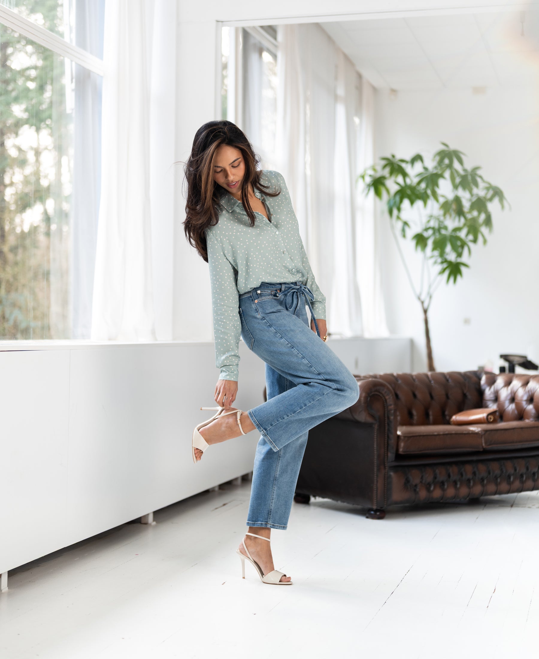 Light stone cropped jeans in denim, modeled indoors near a brown leather sofa and windows.