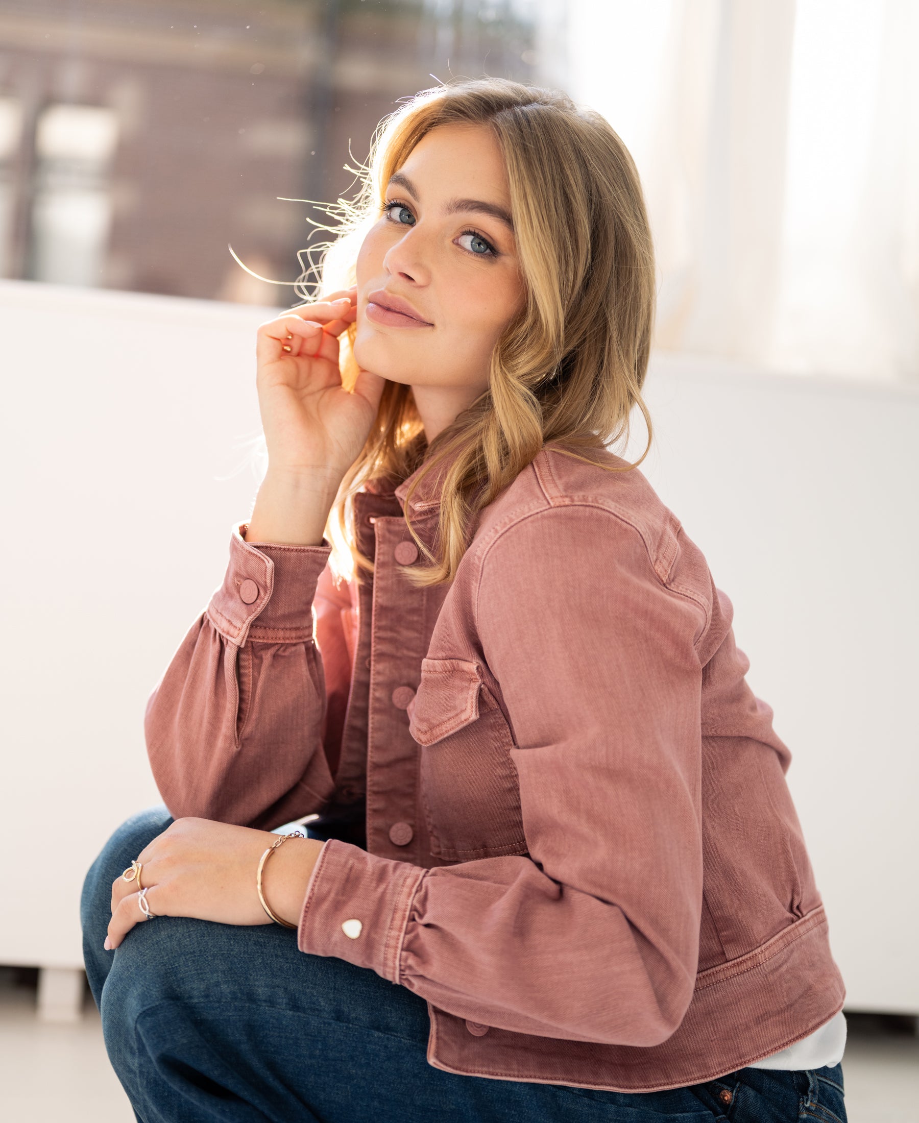 Dusty pink stretch denim jacket, front view indoors. Model is sitting and smiling at the camera.