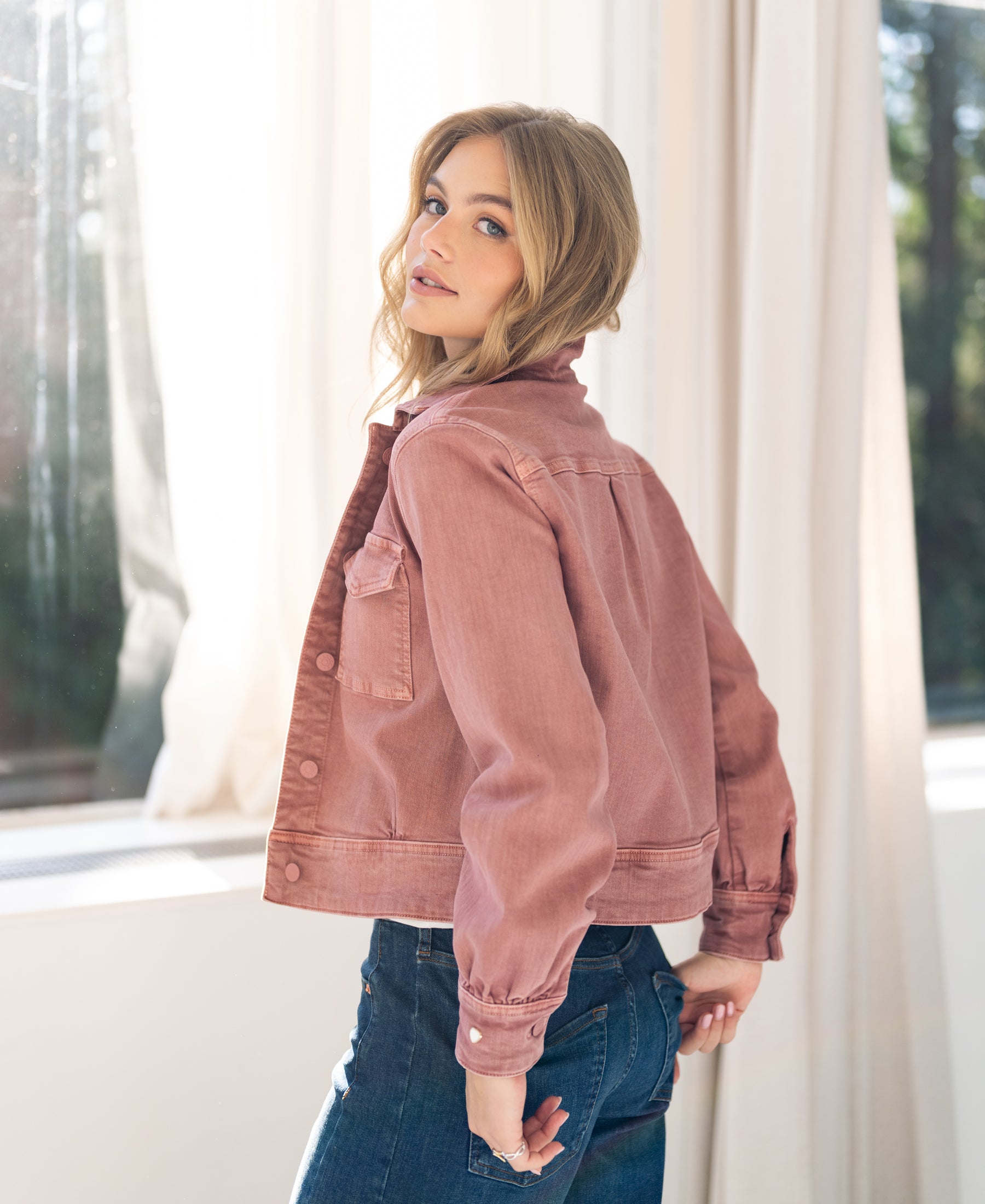 Woman wears dusty pink LA CALLAS stretch denim jacket indoors, looking over her shoulder at the camera.