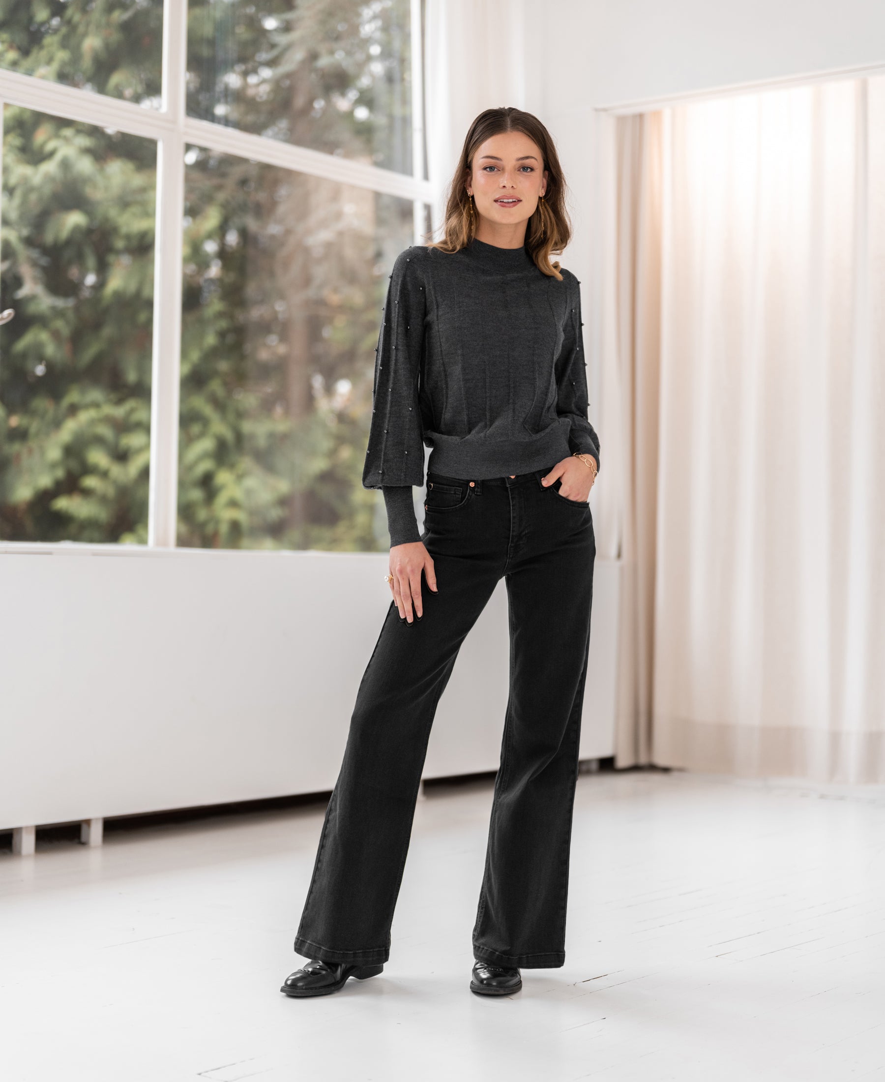 Woman indoors by large windows wearing the Plein Publique beaded charcoal turtleneck sweater.