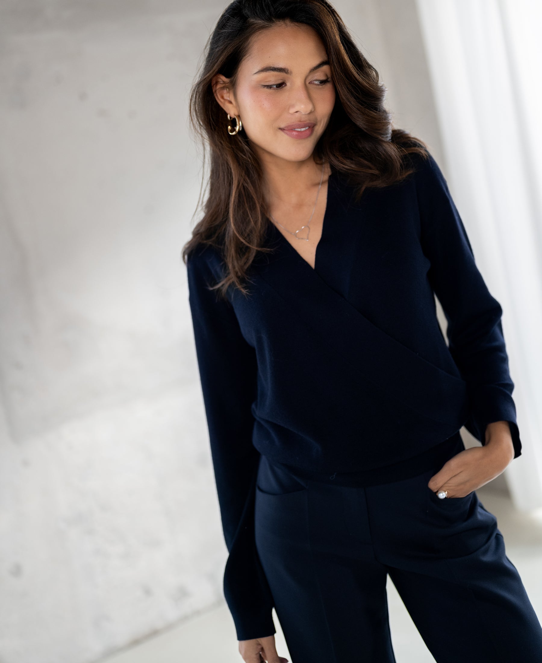 Woman wearing a navy blue LES CINQ wrap sweater by PLEIN PUBLIQUE, standing indoors with one hand in her pocket.