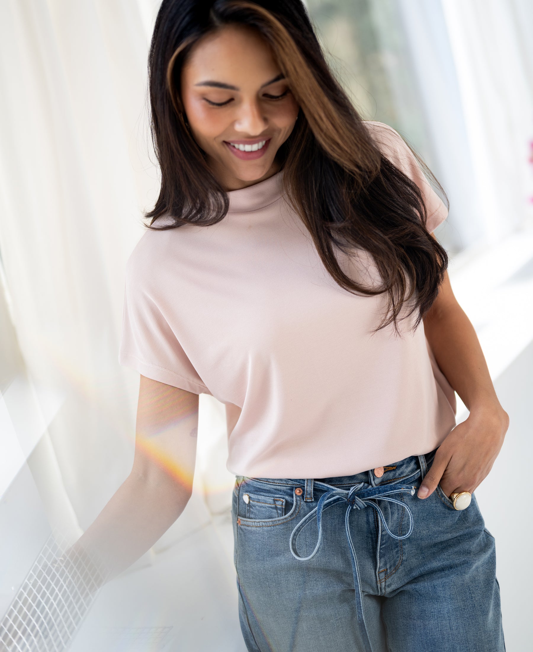Woman wearing a ribbed blush pink collared top stands by a sunlit window, looking down with one hand in her pocket.