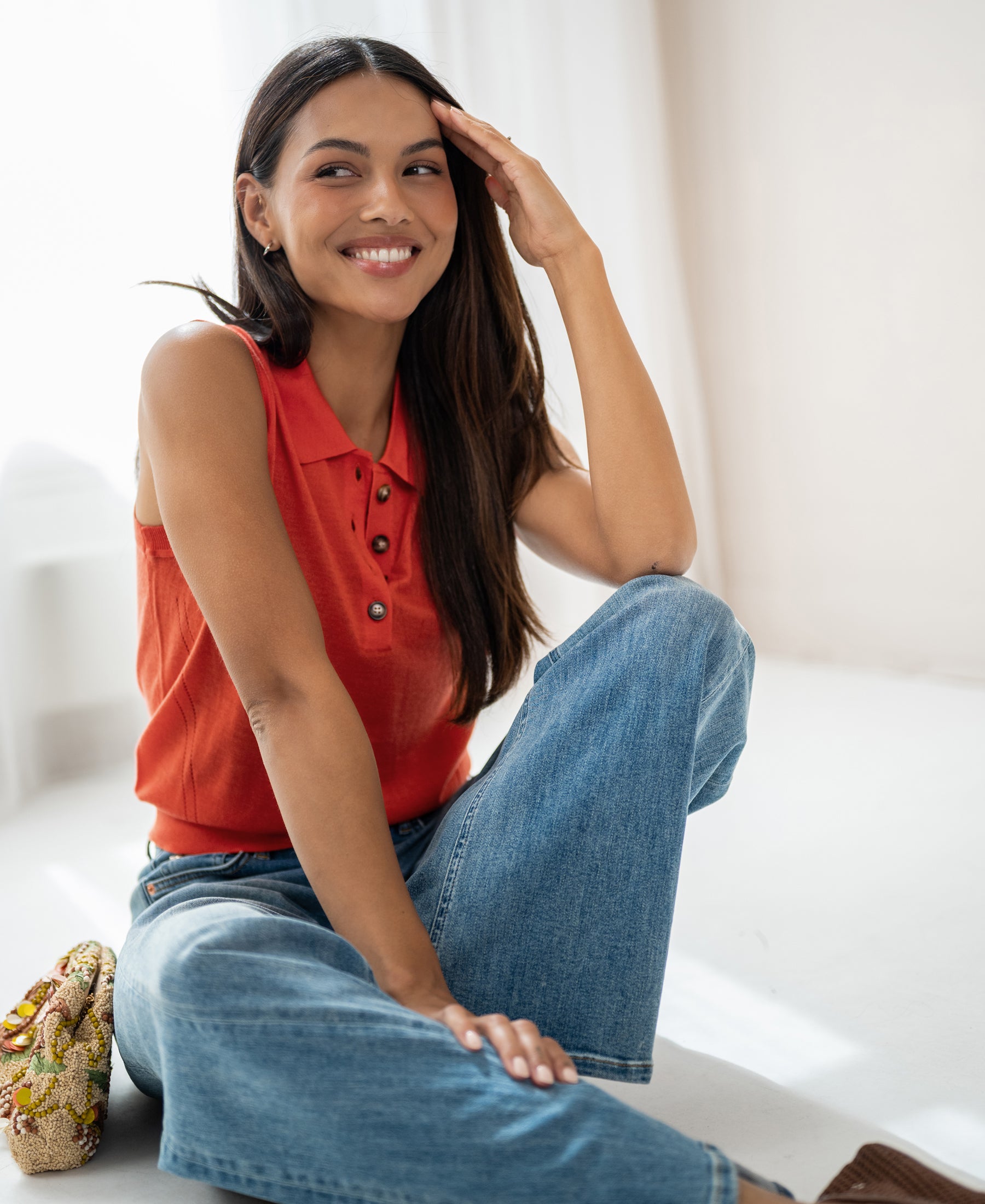 Smiling woman wearing the LE CLUB Coral Sleeveless Polo by PLEIN PUBLIQUE sits on the floor, touching her hair.