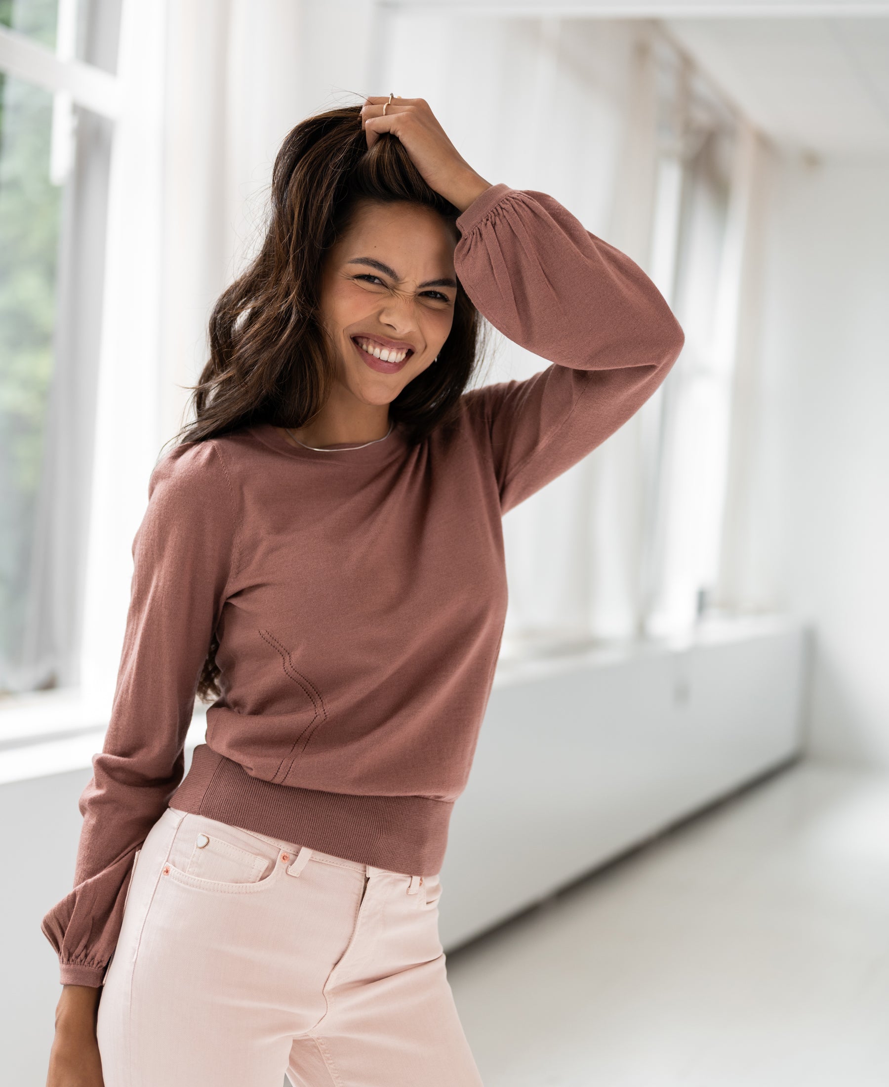 Dusty pink merino wool sweater LA COEUR by PLEIN PUBLIQUE shown on model standing by large windows.