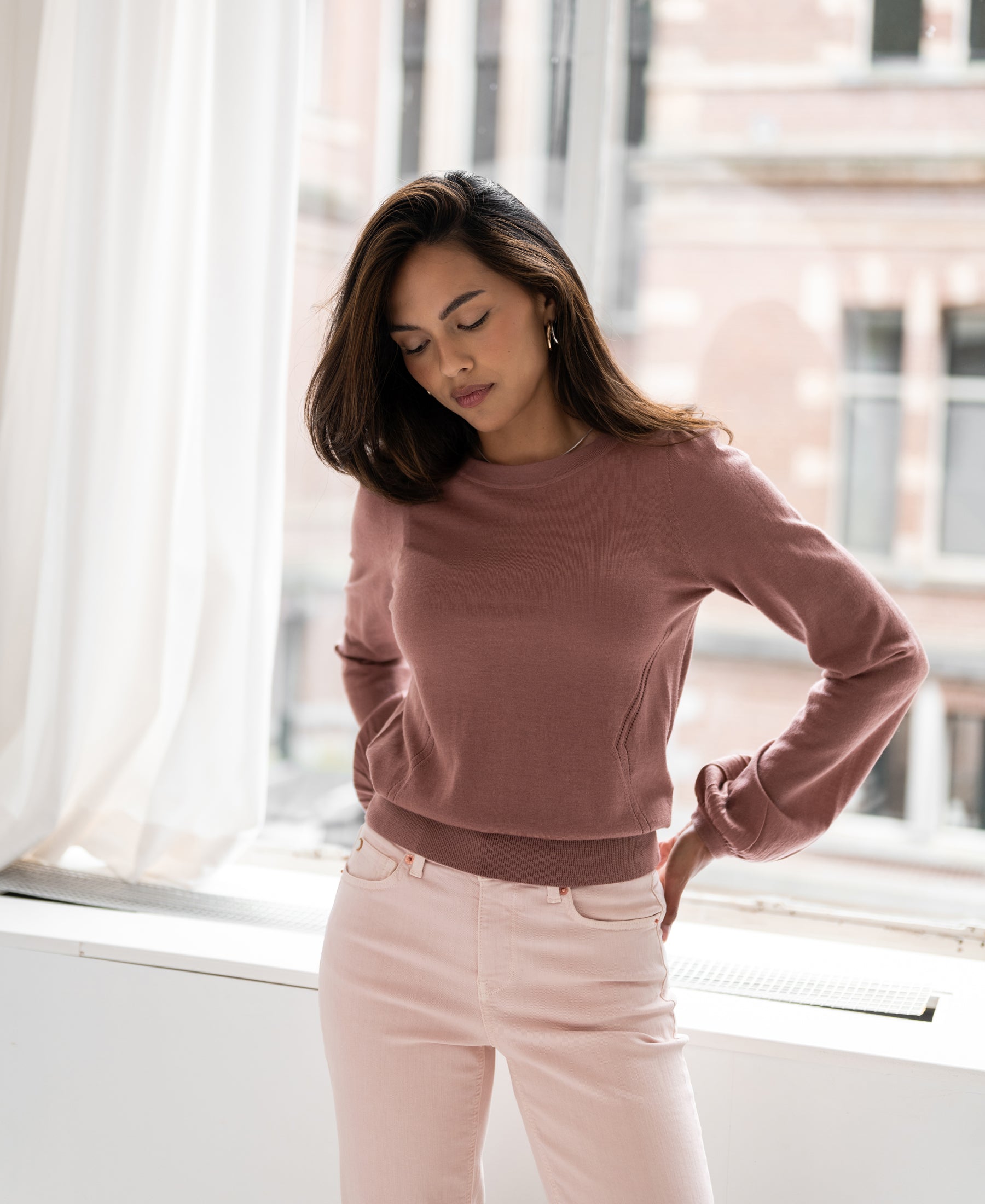Woman wearing a dusty pink merino wool sweater, viewed by a window.