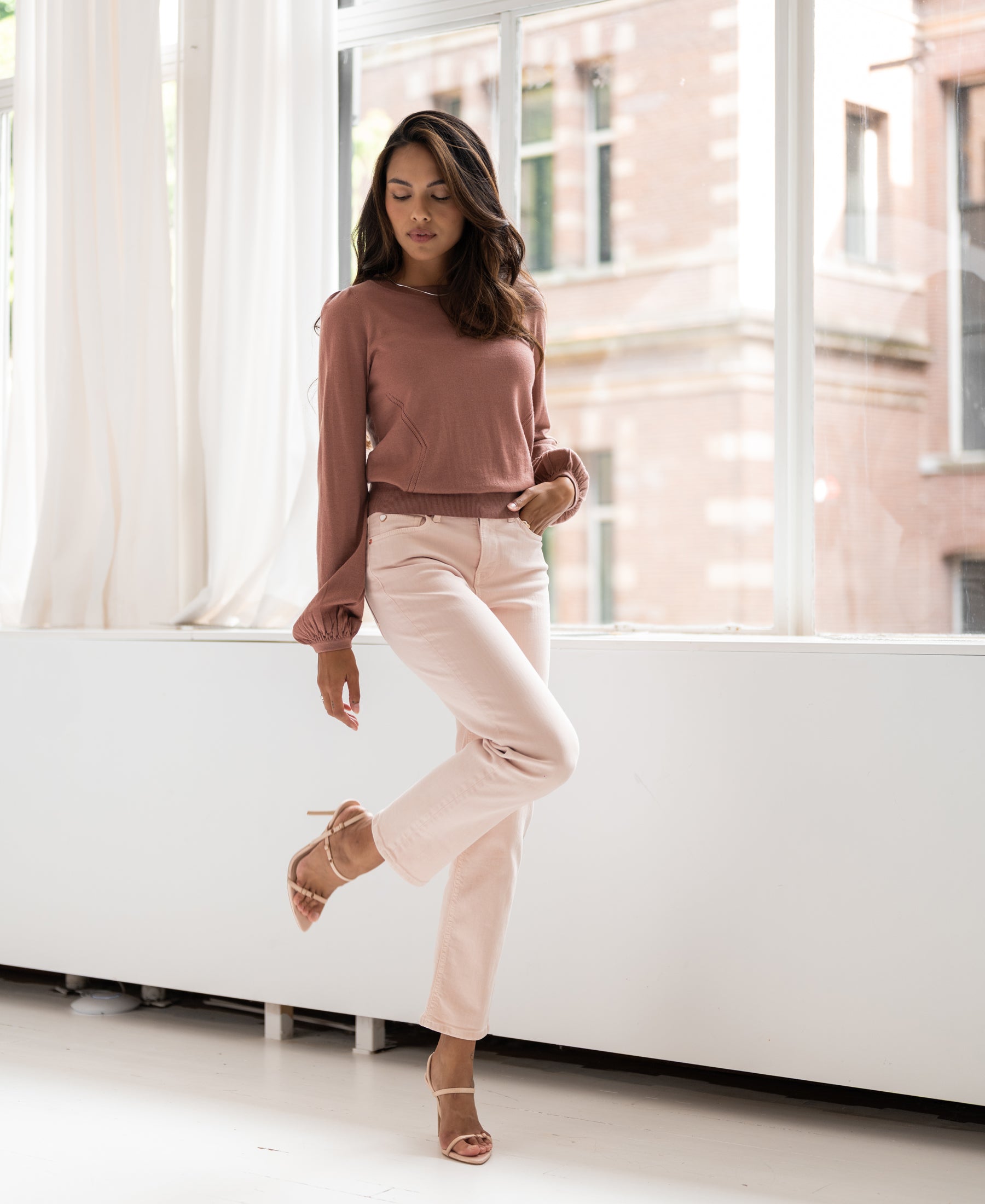 Woman indoors by a window poses in a dusty pink merino wool sweater, one leg raised.