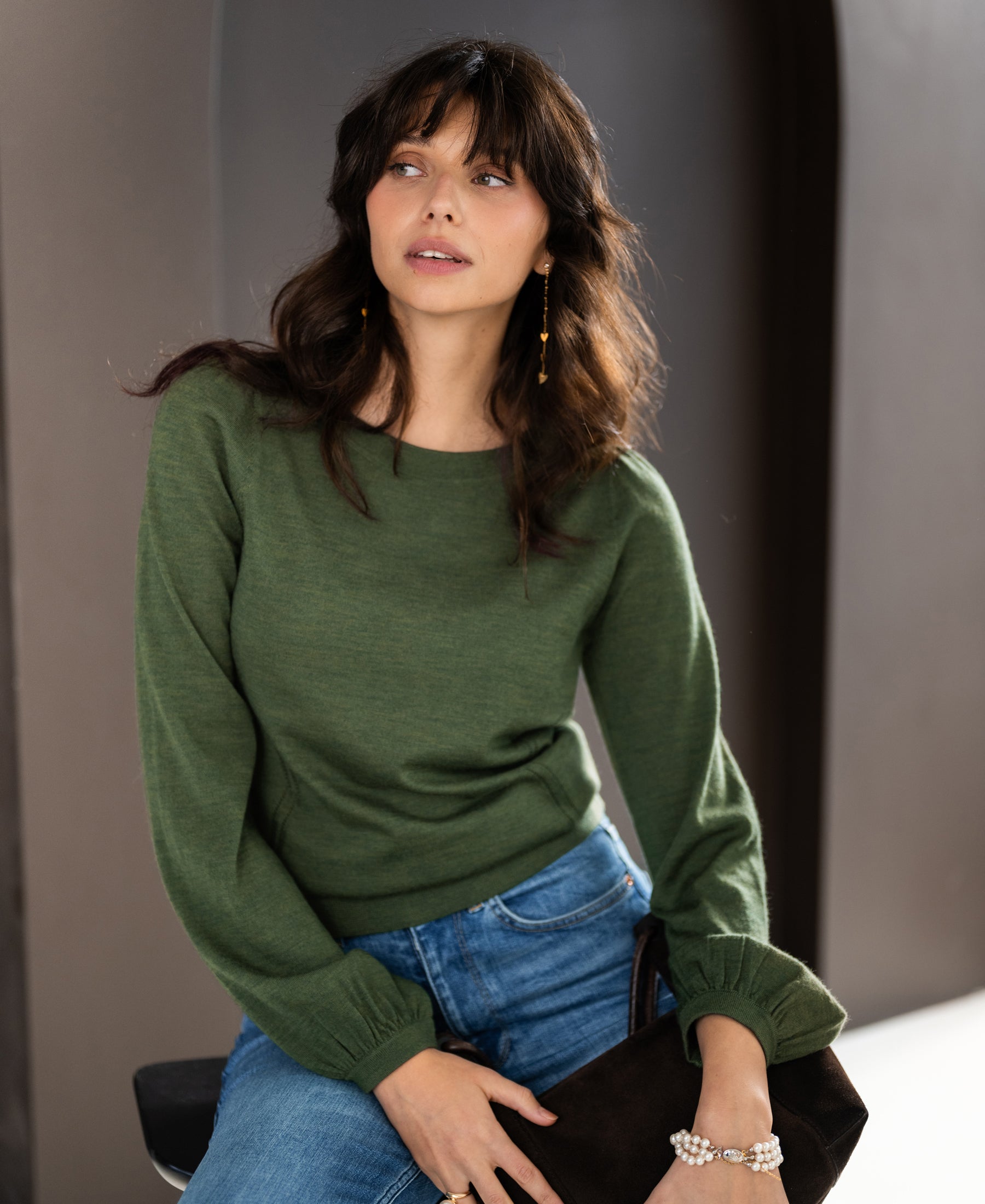 Woman with wavy brown hair wearing an olive green Merino wool sweater called LA COEUR by PLEIN PUBLIQUE.