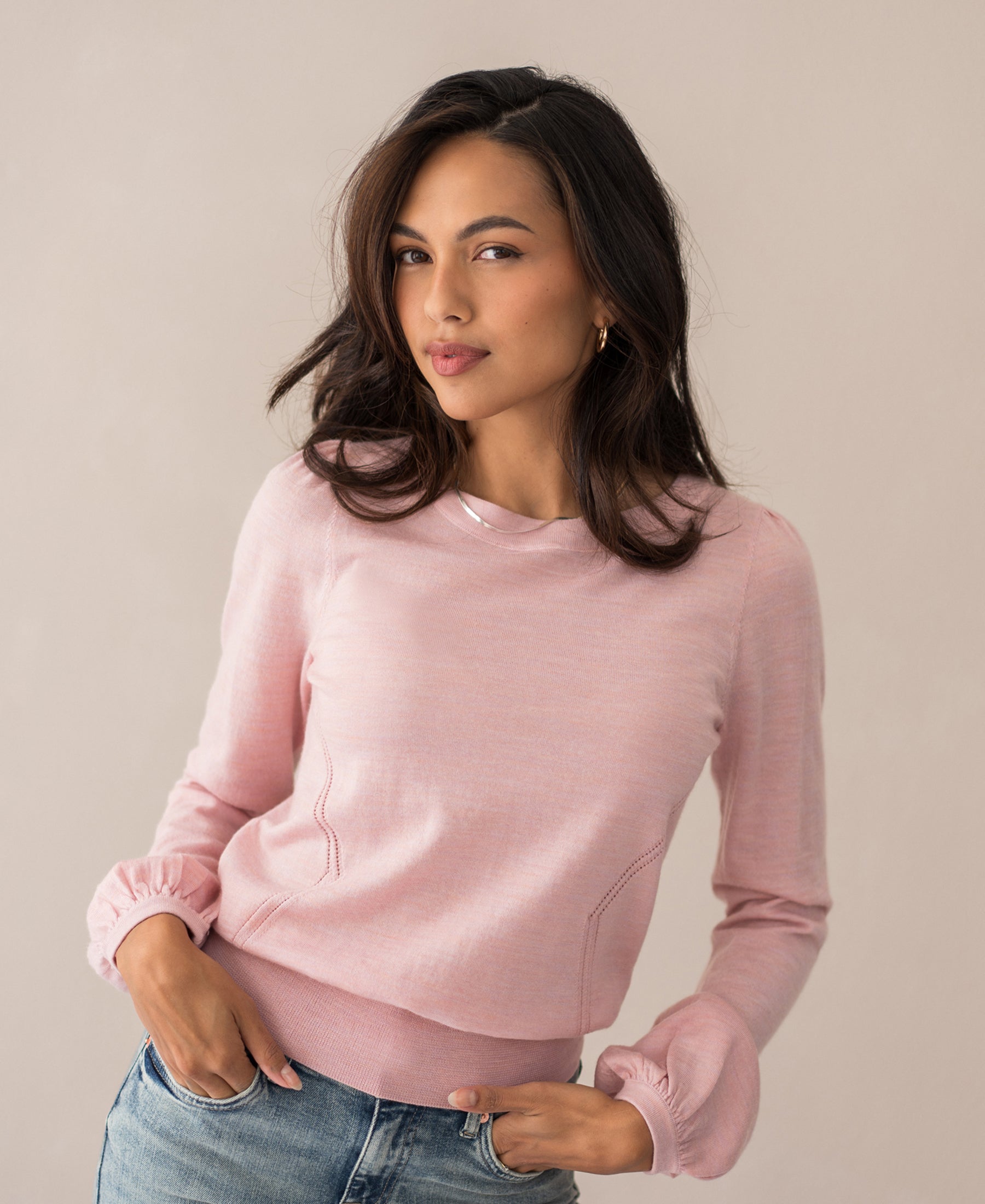 A woman with long dark hair wears the Merinotrui LA COEUR Rose—a light pink merino wool sweater with ajour details—paired with blue jeans, standing against a neutral background and looking confidently at the camera.