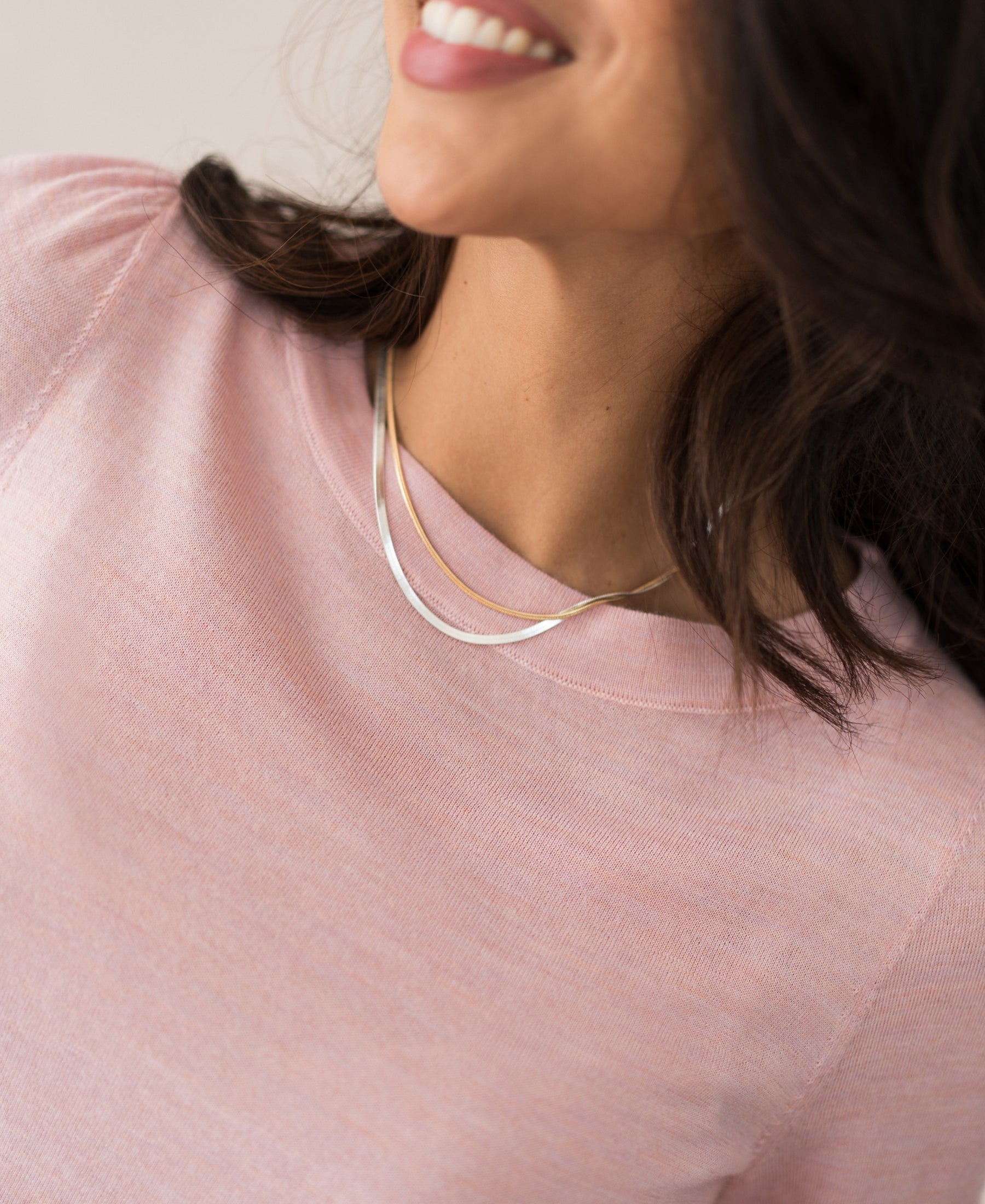 A woman with wavy dark hair smiles, wearing the Merinotrui LA COEUR Rose—a light pink top with balloon sleeves—and two delicate layered gold necklaces.