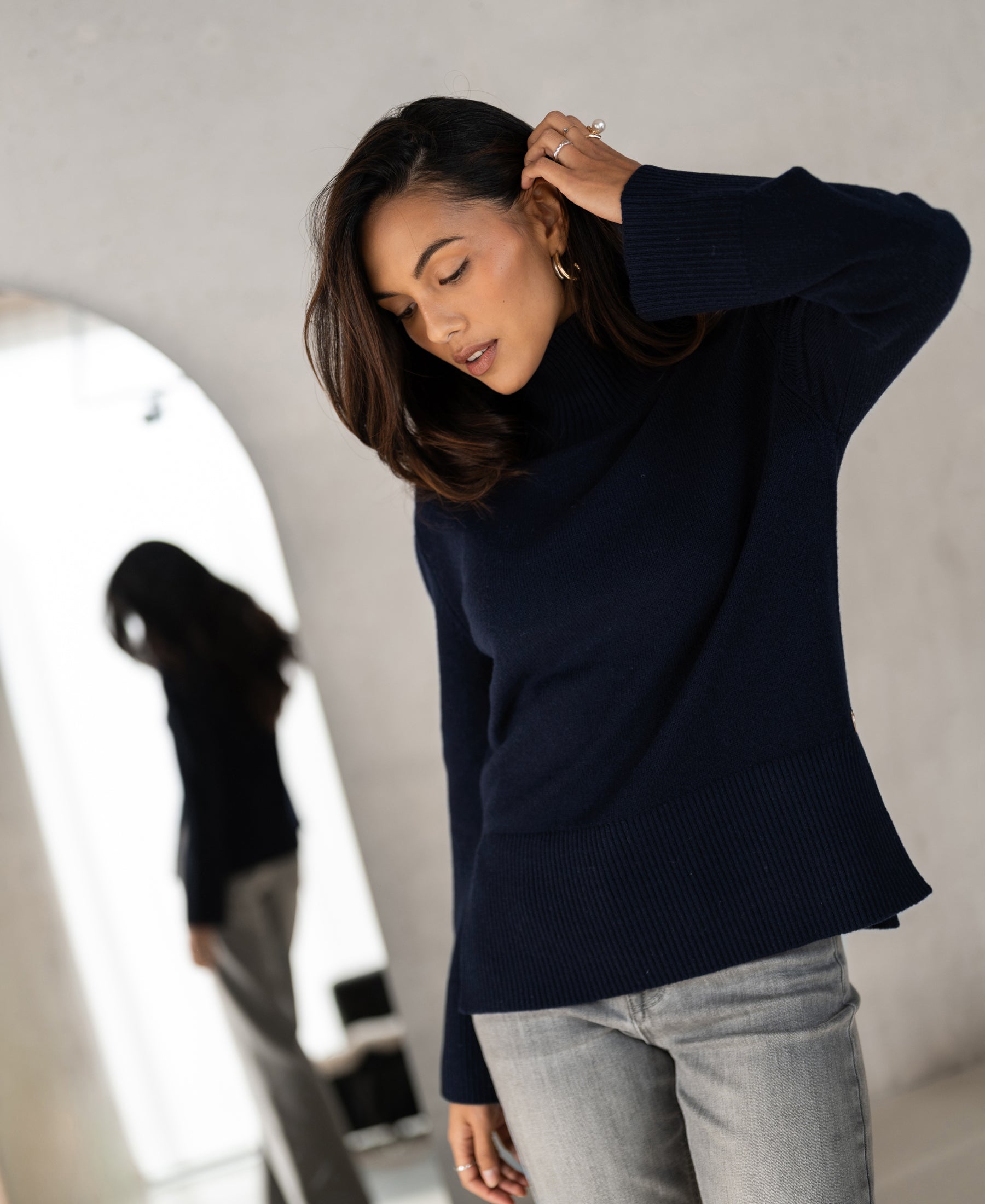 Woman wearing a PLEIN PUBLIQUE LA COZY dark blue turtleneck sweater stands indoors by a mirror.