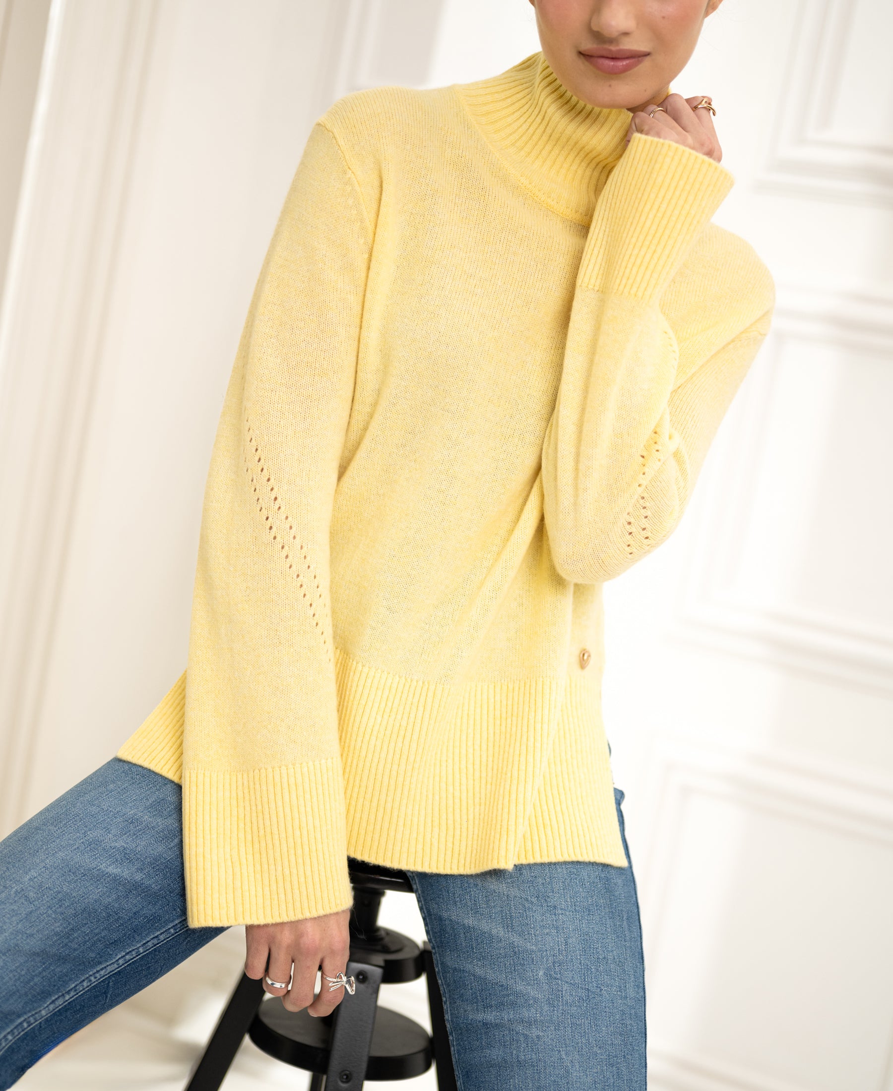 Woman wearing a yellow PLEIN PUBLIQUE LA COZY turtleneck sweater, seated against a white background.