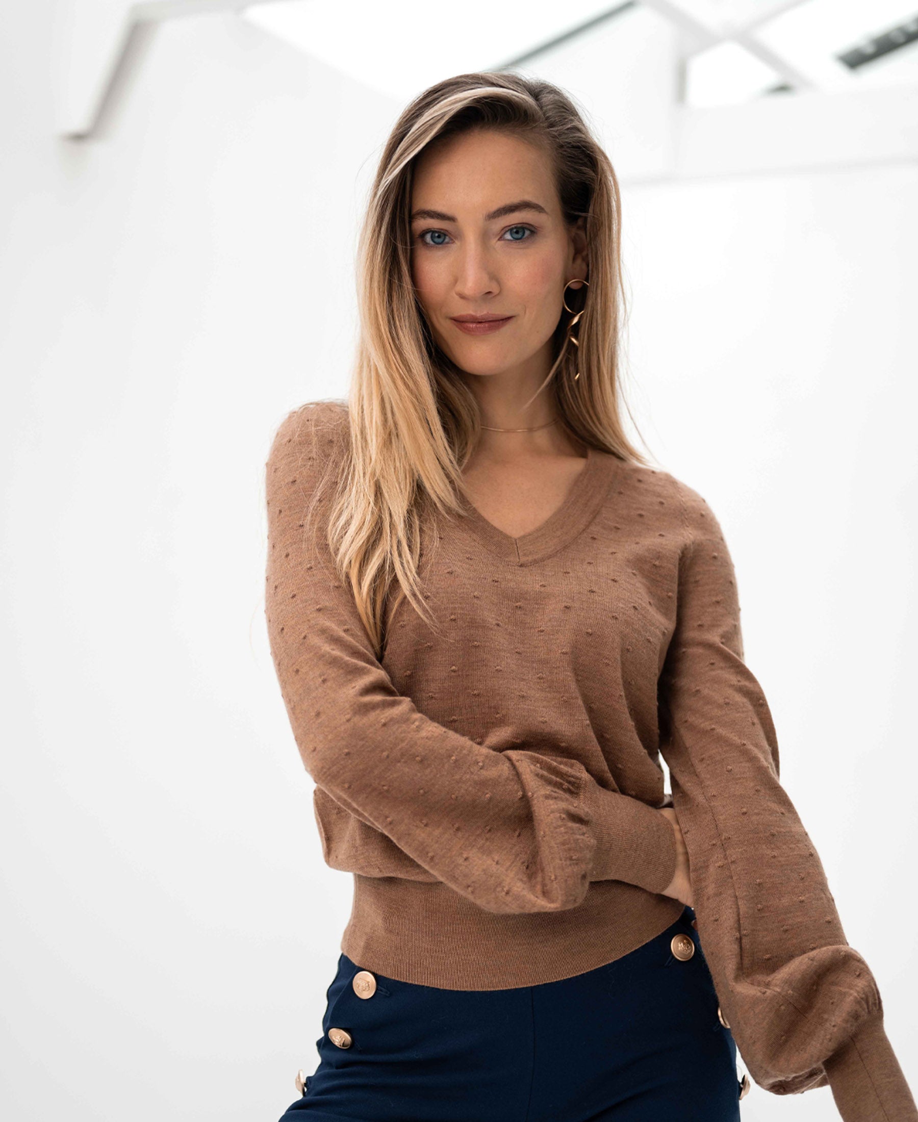 PLEIN PUBLIQUE Merino wool V-neck sweater LA VICTORIA in Chai Tea, worn by a woman with long blond hair in a bright room.