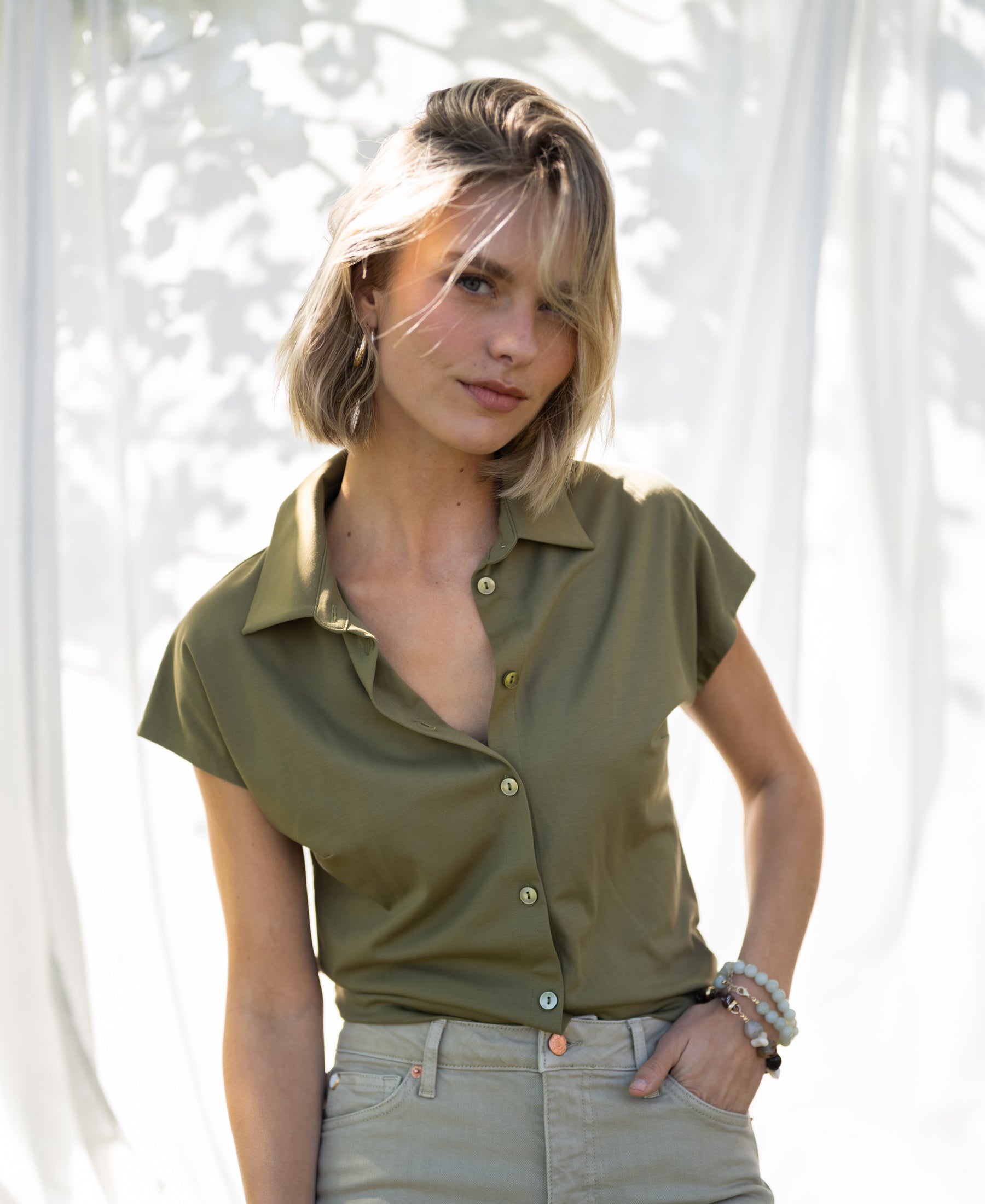 Olive PLEIN PUBLIQUE travel blouse LA DELON shown outdoors with sunlight behind, one hand in pocket.