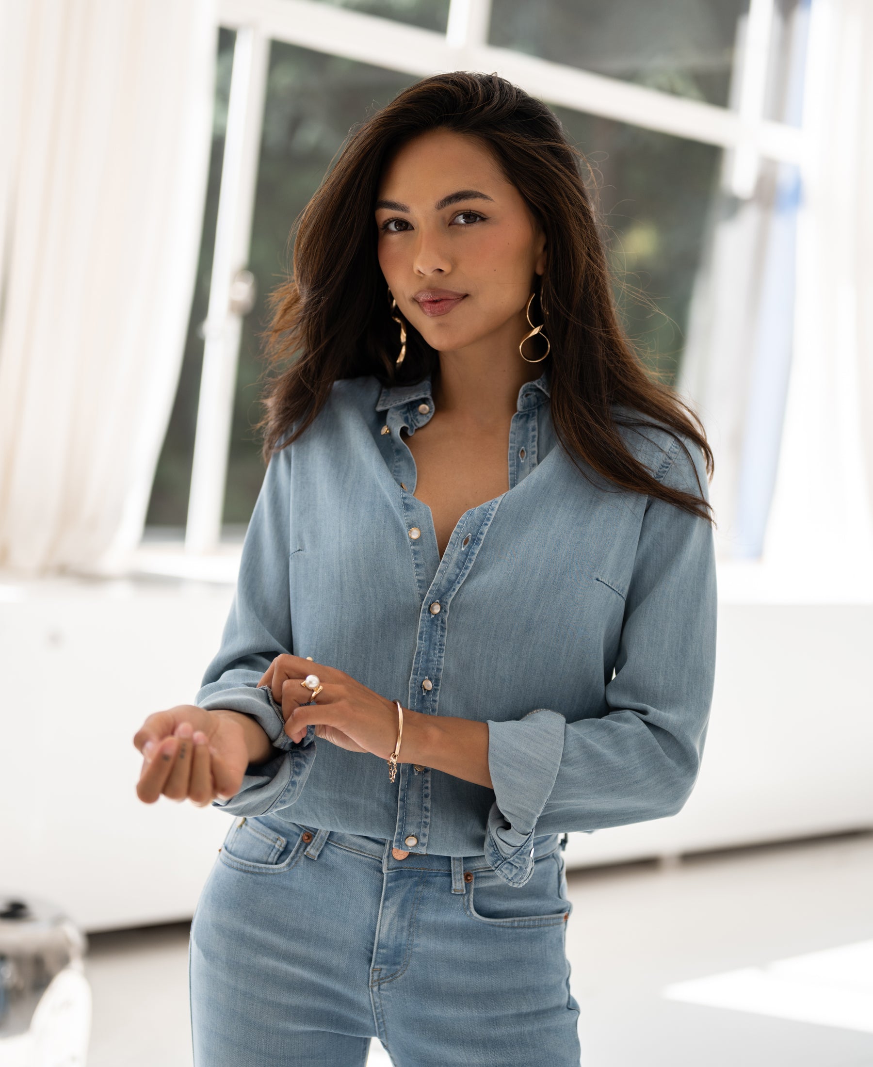 Woman wearing a bleached Tencel denim blouse, standing by white curtains.