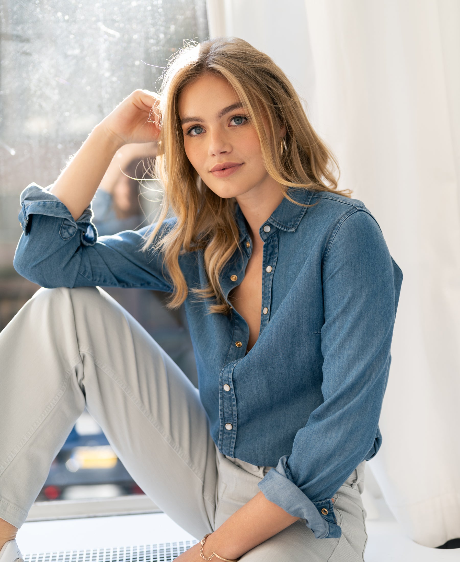 Woman wearing a stonewash Tencel denim blouse sits by a window, smiling at the camera.