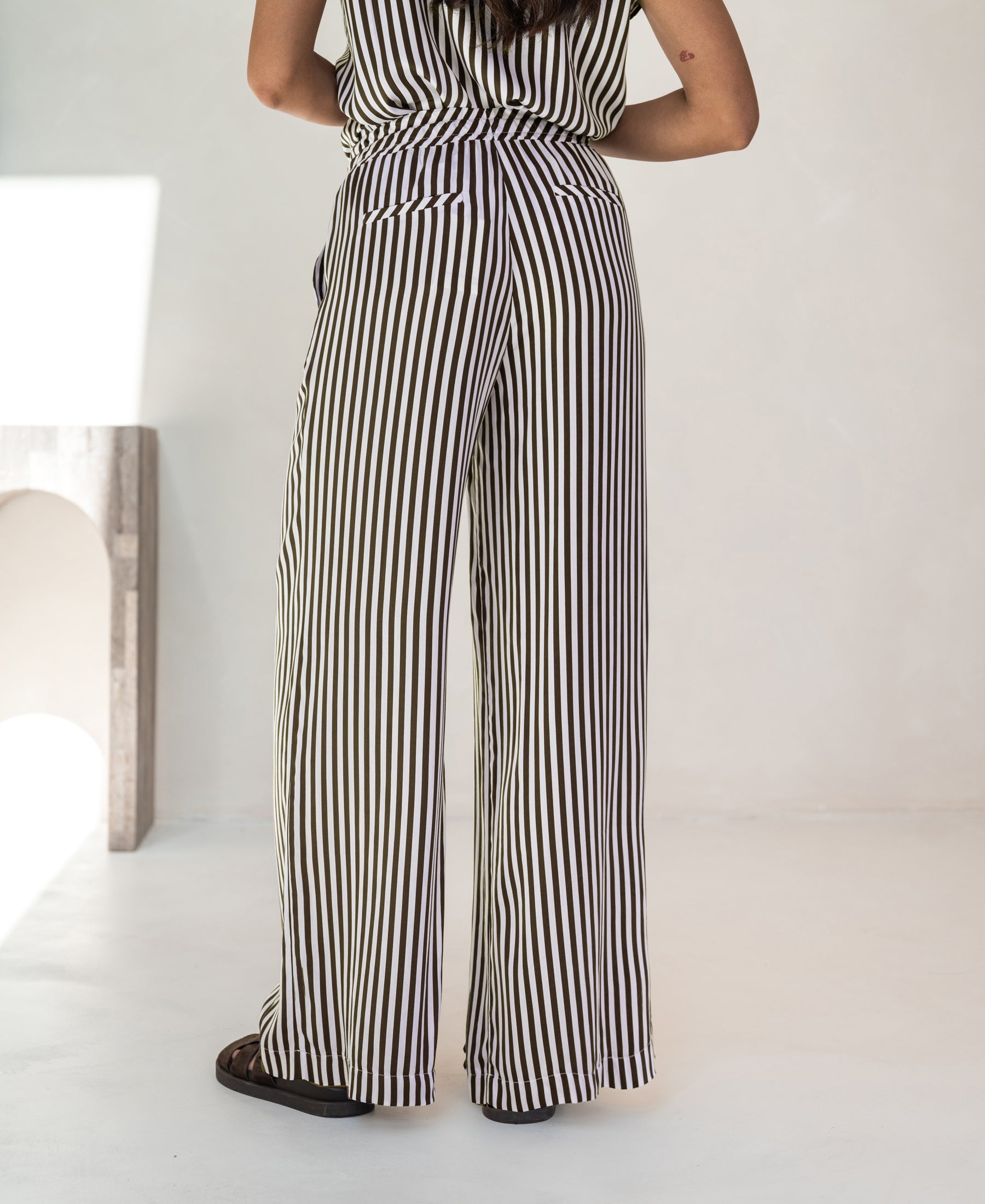 Woman wearing ivory-cocoa striped summer trousers, shown from the back, indoors.