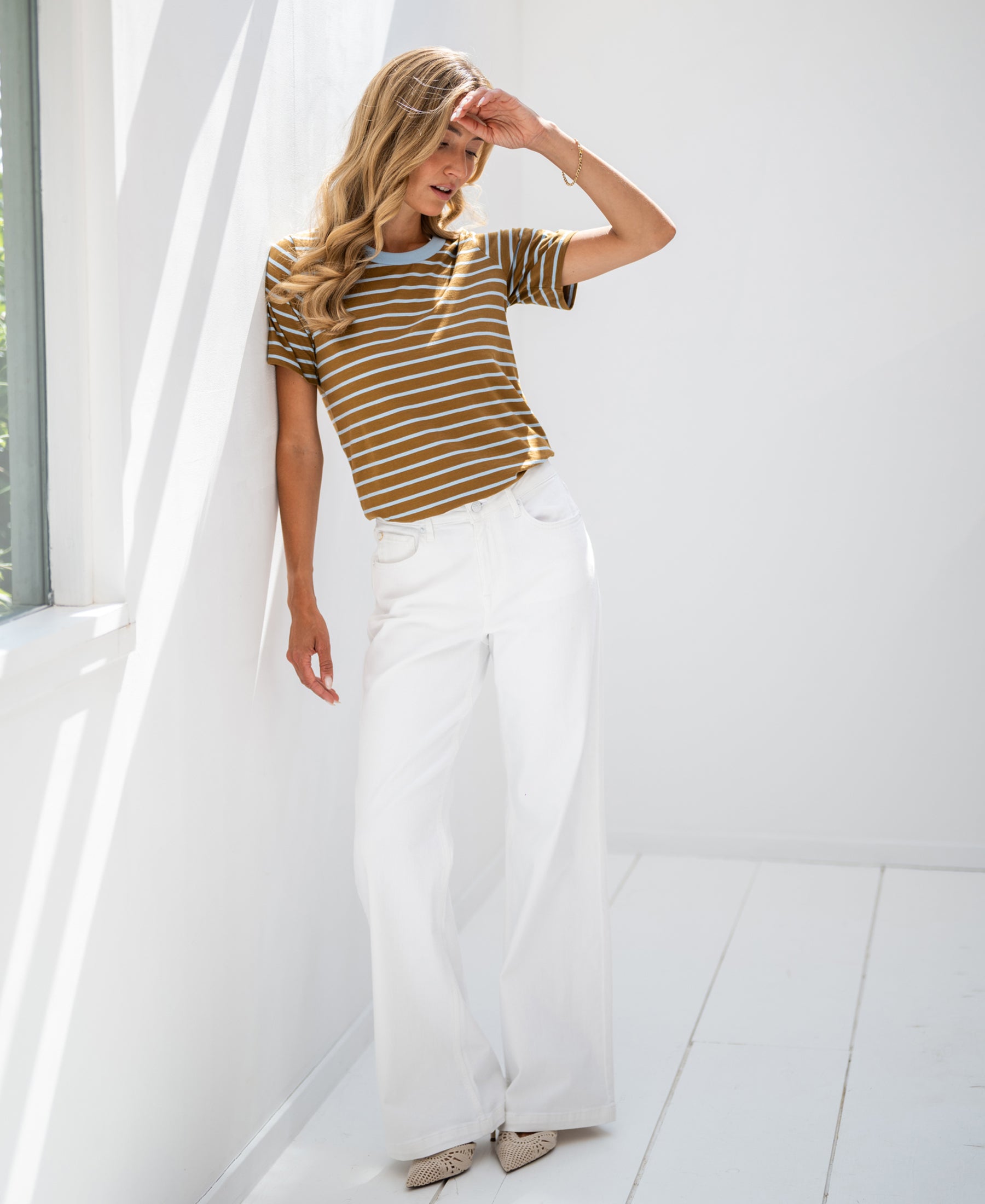 Woman wearing a striped LA DYLAN T-shirt stands against a sunlit wall, shielding her eyes from the sunlight.