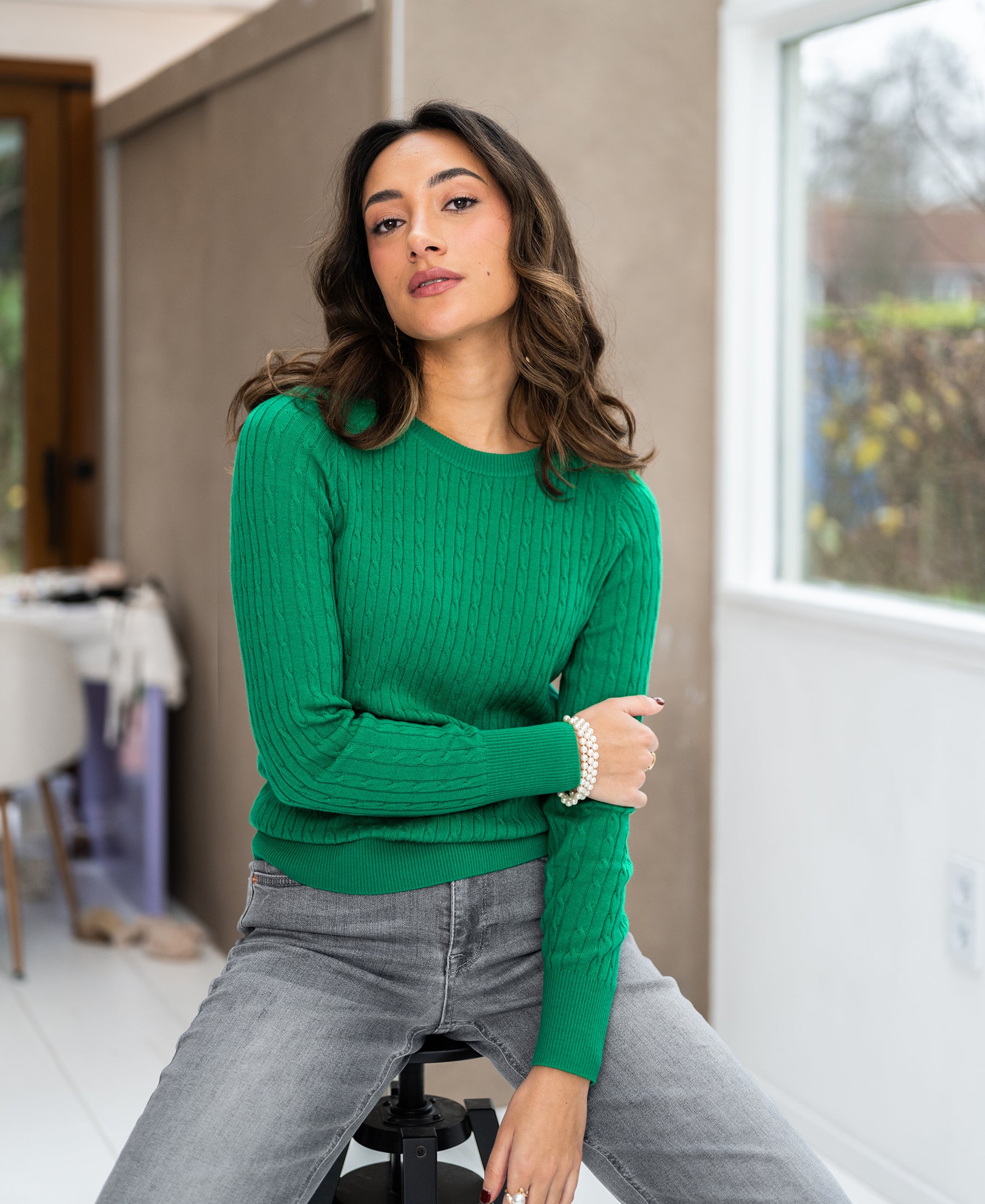 Woman wearing bright green ETHAN cable-knit sweater sitting indoors on a stool.