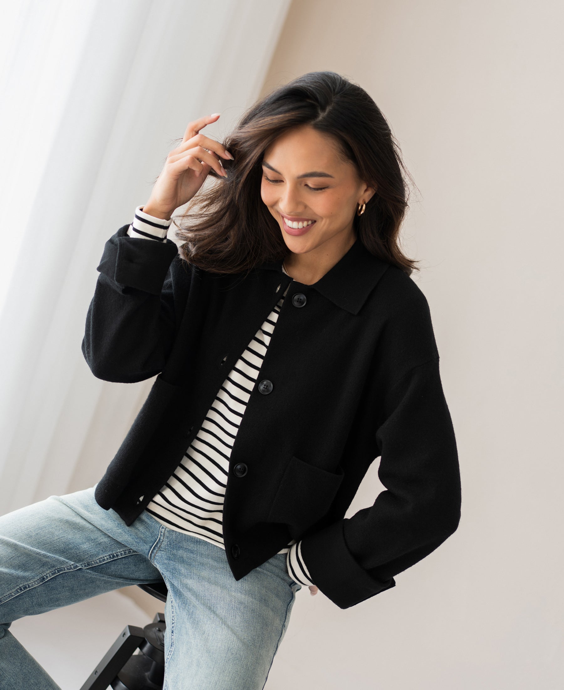 Front view of woman wearing a black wool jacket, seated on a stool with one hand in her hair.