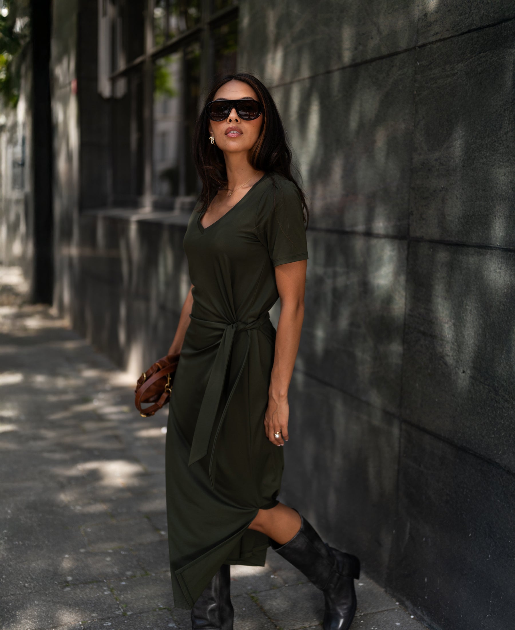 Woman wears a dark olive LA FILICIA long dress by PLEIN PUBLIQUE, walking by a dark wall.