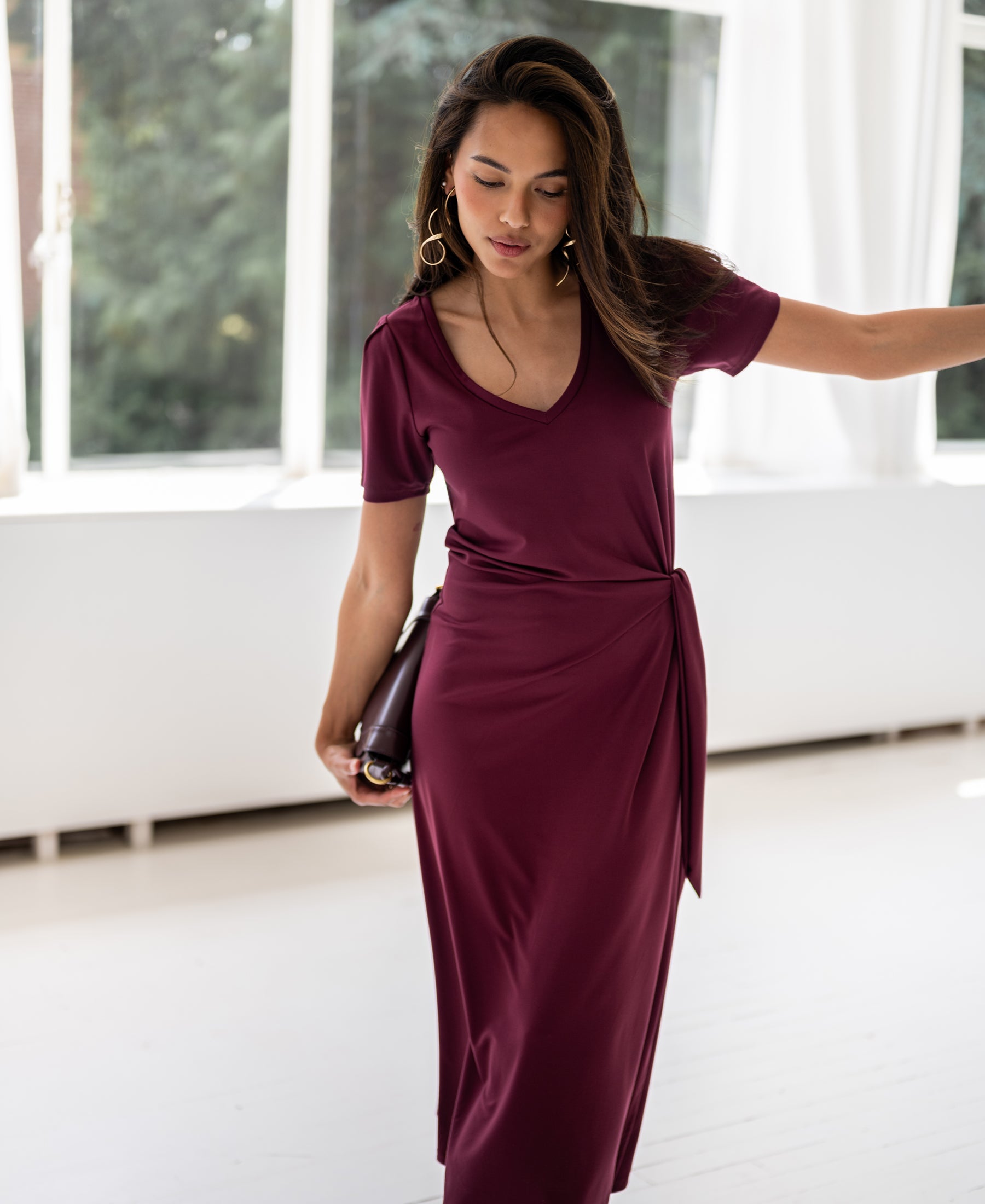 Woman wearing a long wine-colored dress stands indoors by windows, looking down.