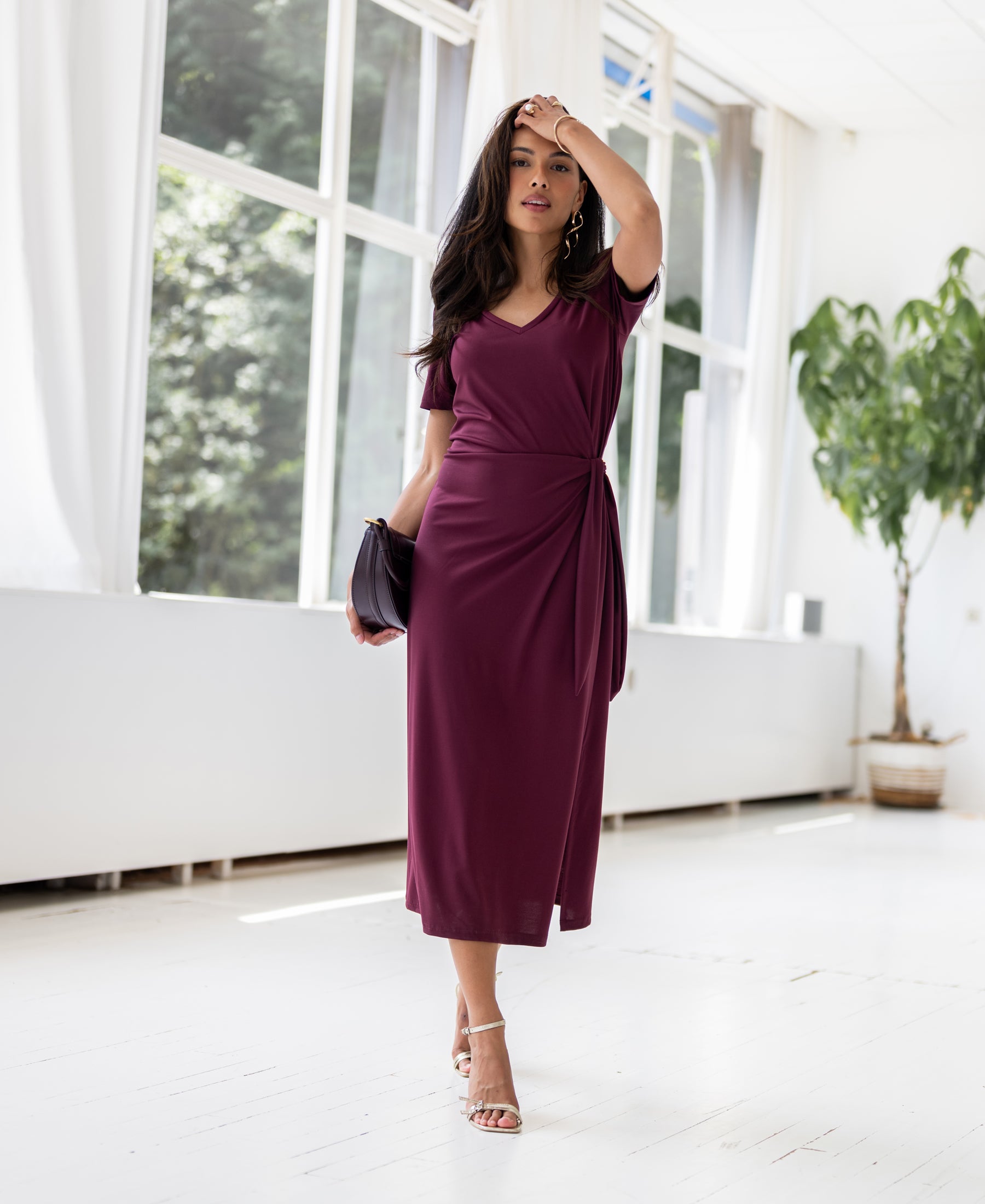 Wine red long dress LA FILICIA by PLEIN PUBLIQUE shown in a bright room with large windows and a potted plant.
