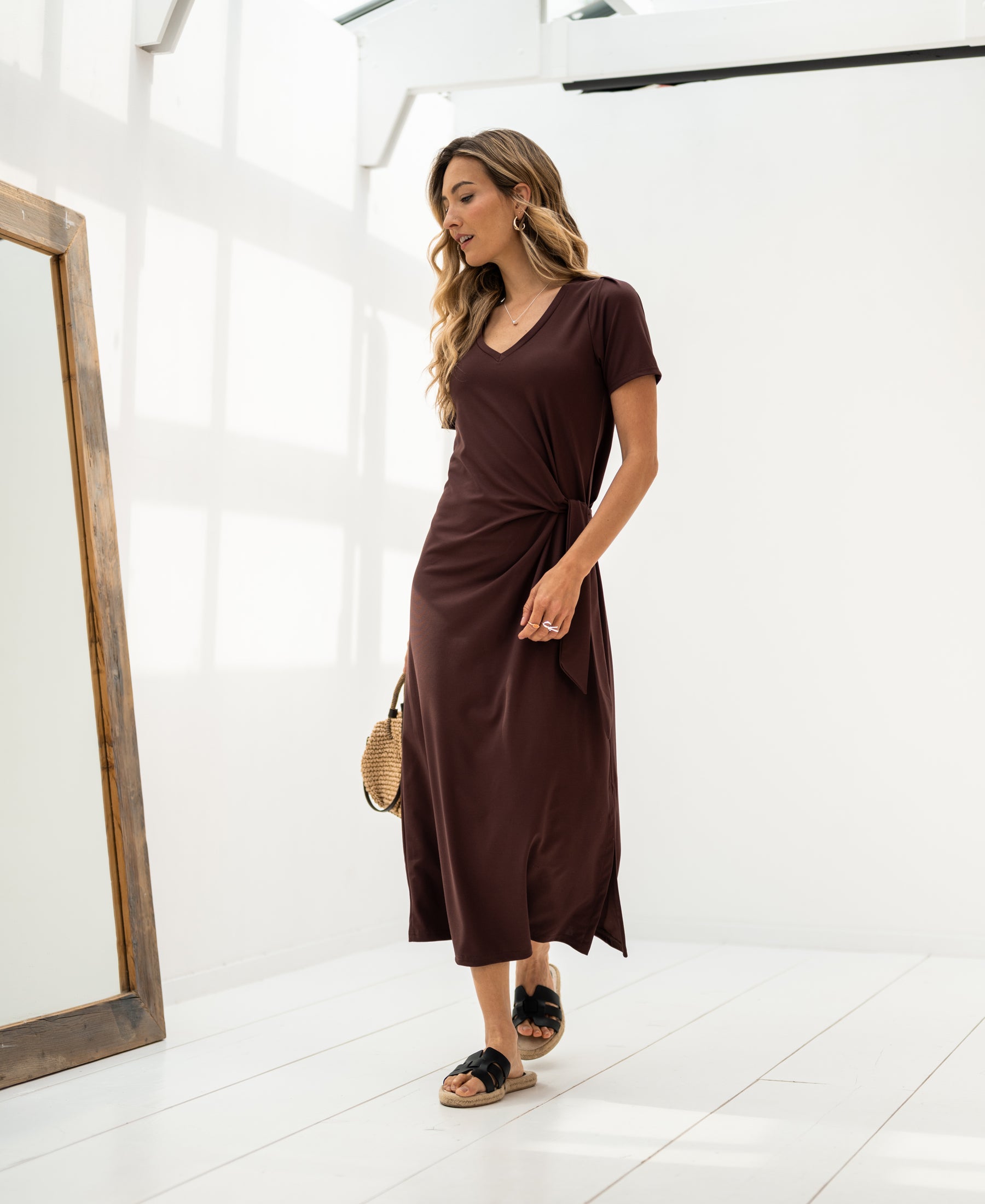 Woman wearing a long dress, LA FILICIA Cacao by PLEIN PUBLIQUE, stands indoors beside a large mirror.