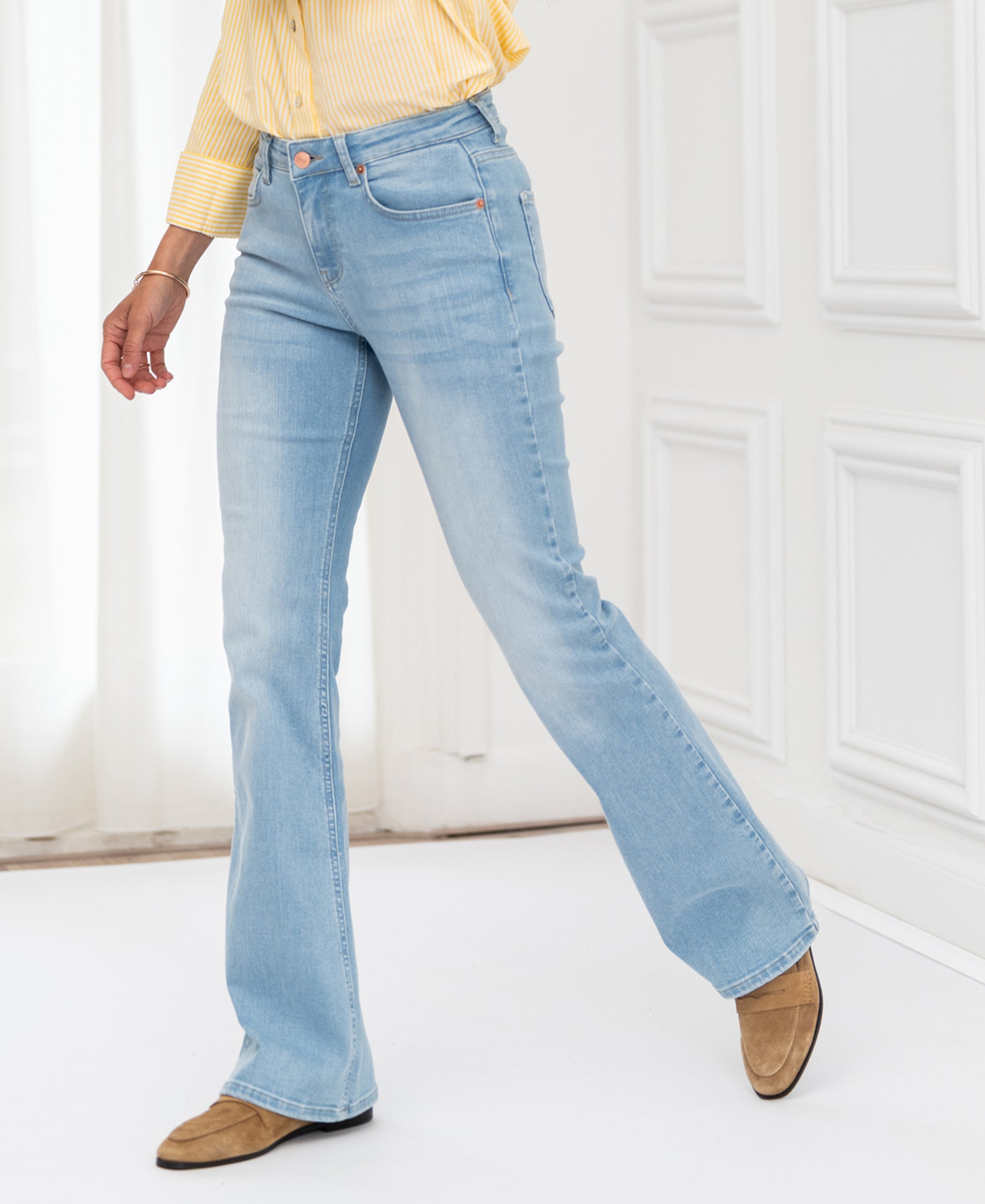 Person wearing bleached flared jeans stands indoors by white curtains.