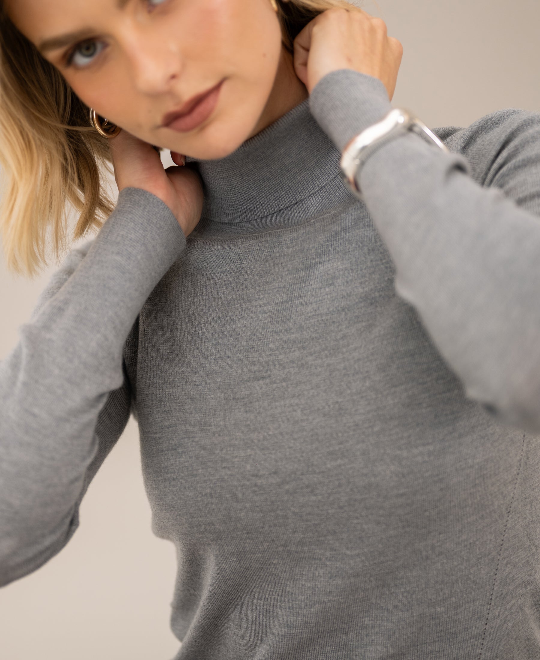 Woman wearing merino wool turtleneck sweater in feather grey, adjusting collar, against neutral background.
