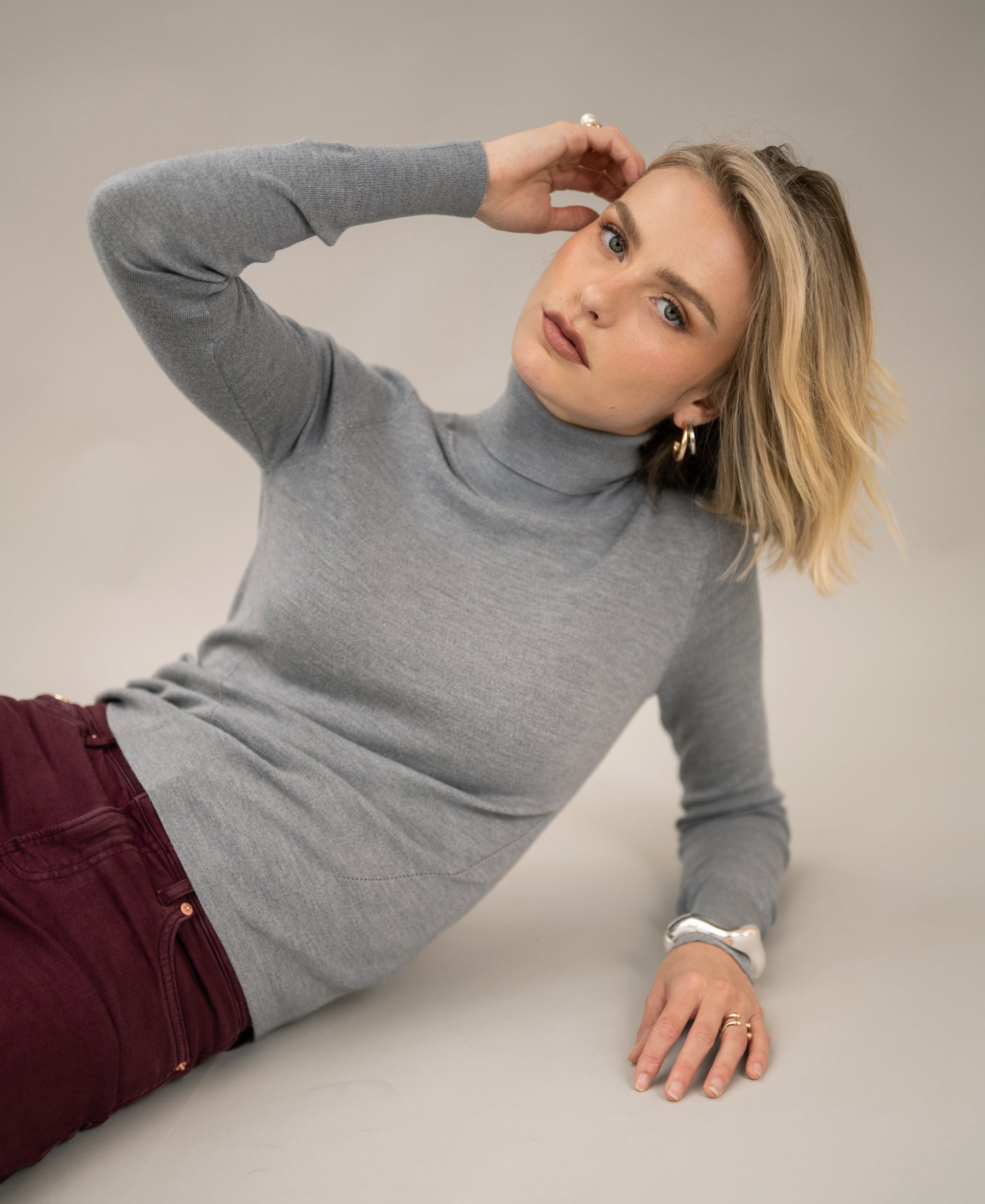 Feathergrey Merino wool turtleneck sweater by PLEIN PUBLIQUE, shown on a model reclining and looking at the camera.