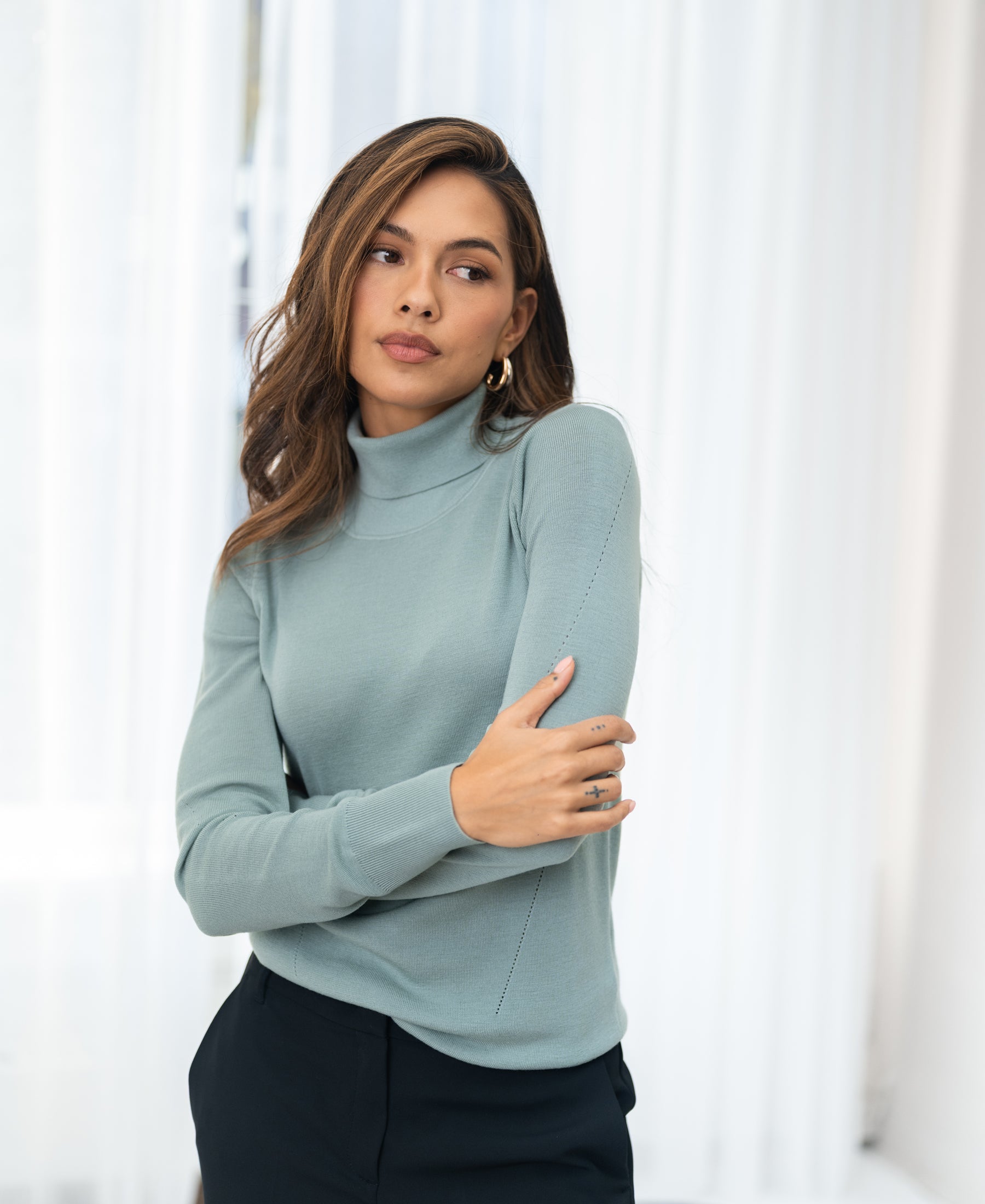 Woman indoors in the PLEIN PUBLIQUE LA FORTE Sage Merino turtleneck sweater, looking to the side.