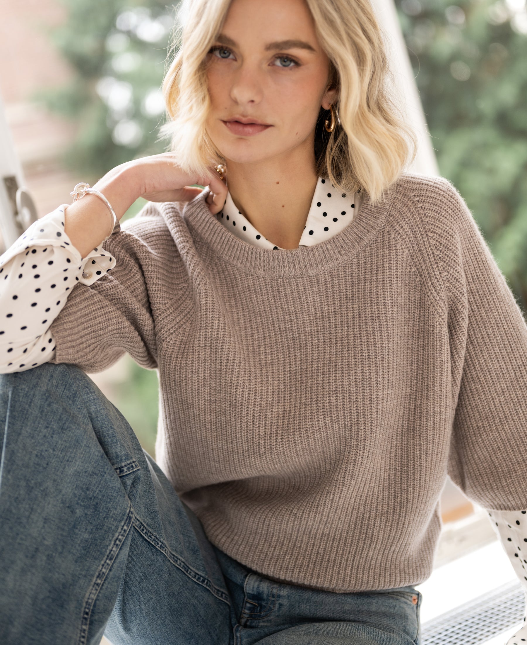 Warm taupe sweater by PLEIN PUBLIQUE, Trui LA FRAMBOISE, shown indoors. Green trees visible outside the window.