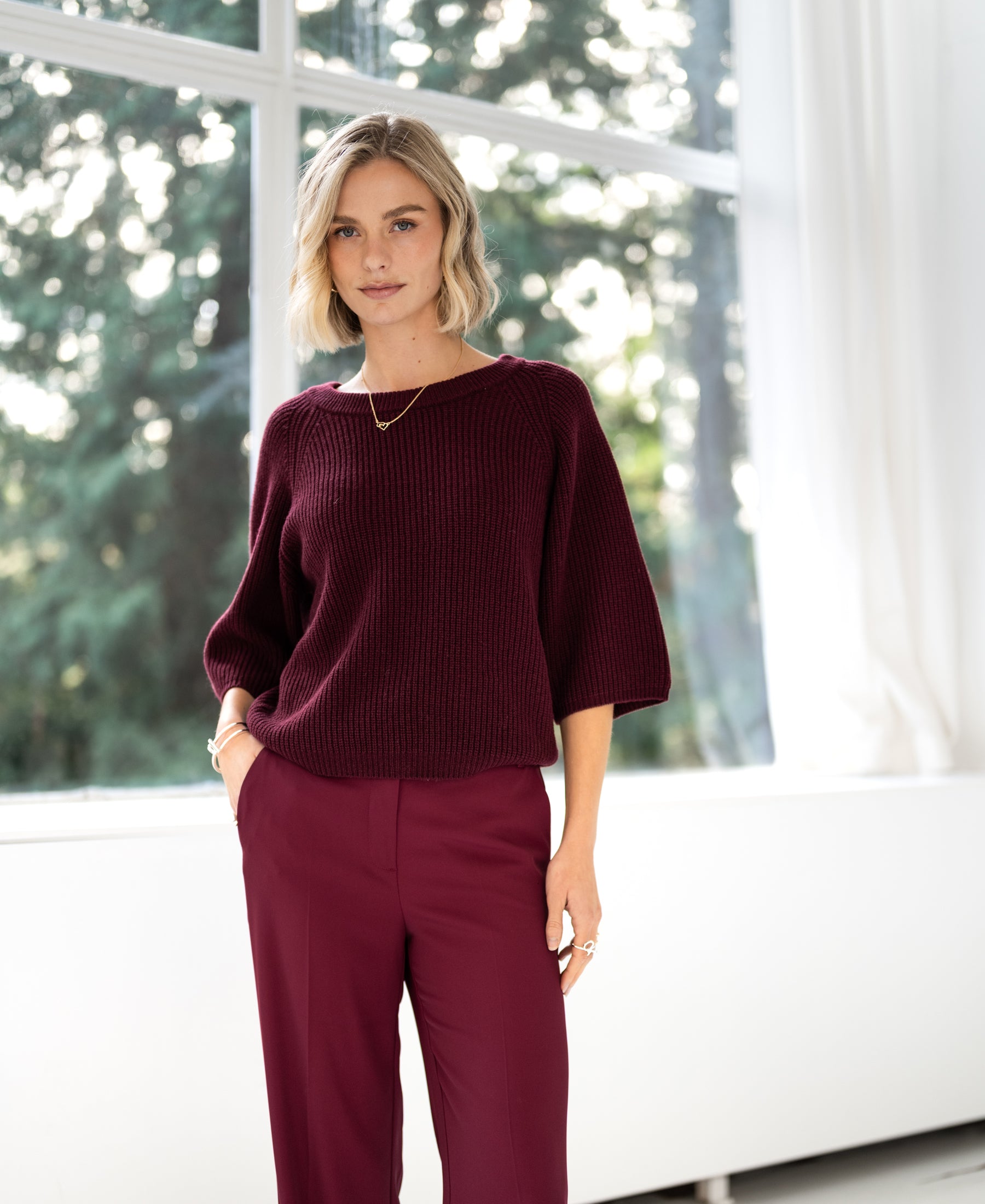 Woman wearing a LA FRAMBOISE Wine Red Sweater by PLEIN PUBLIQUE stands indoors near a window with trees outside.