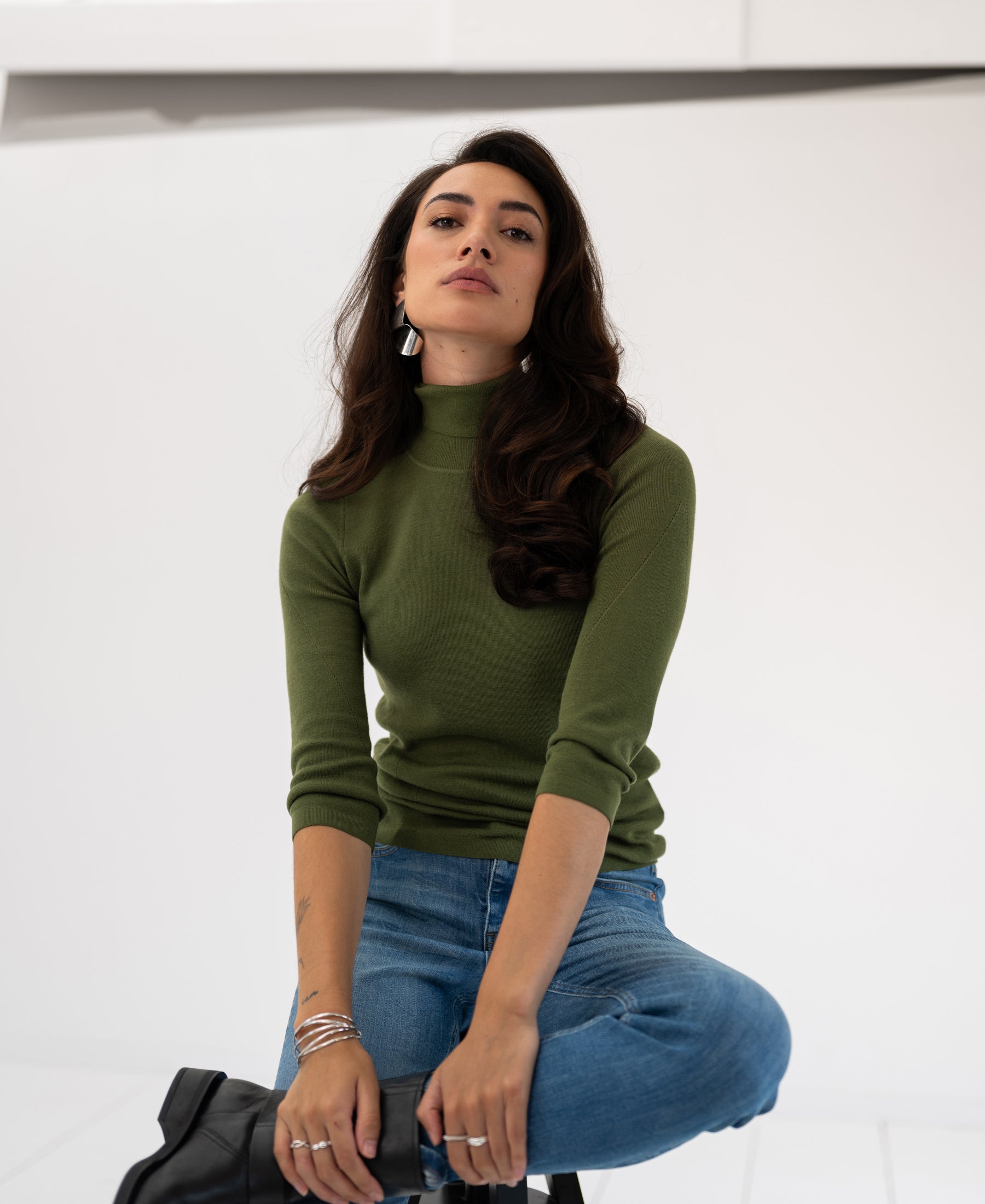 Woman wearing an olive green turtleneck sweater, seated against a white background.