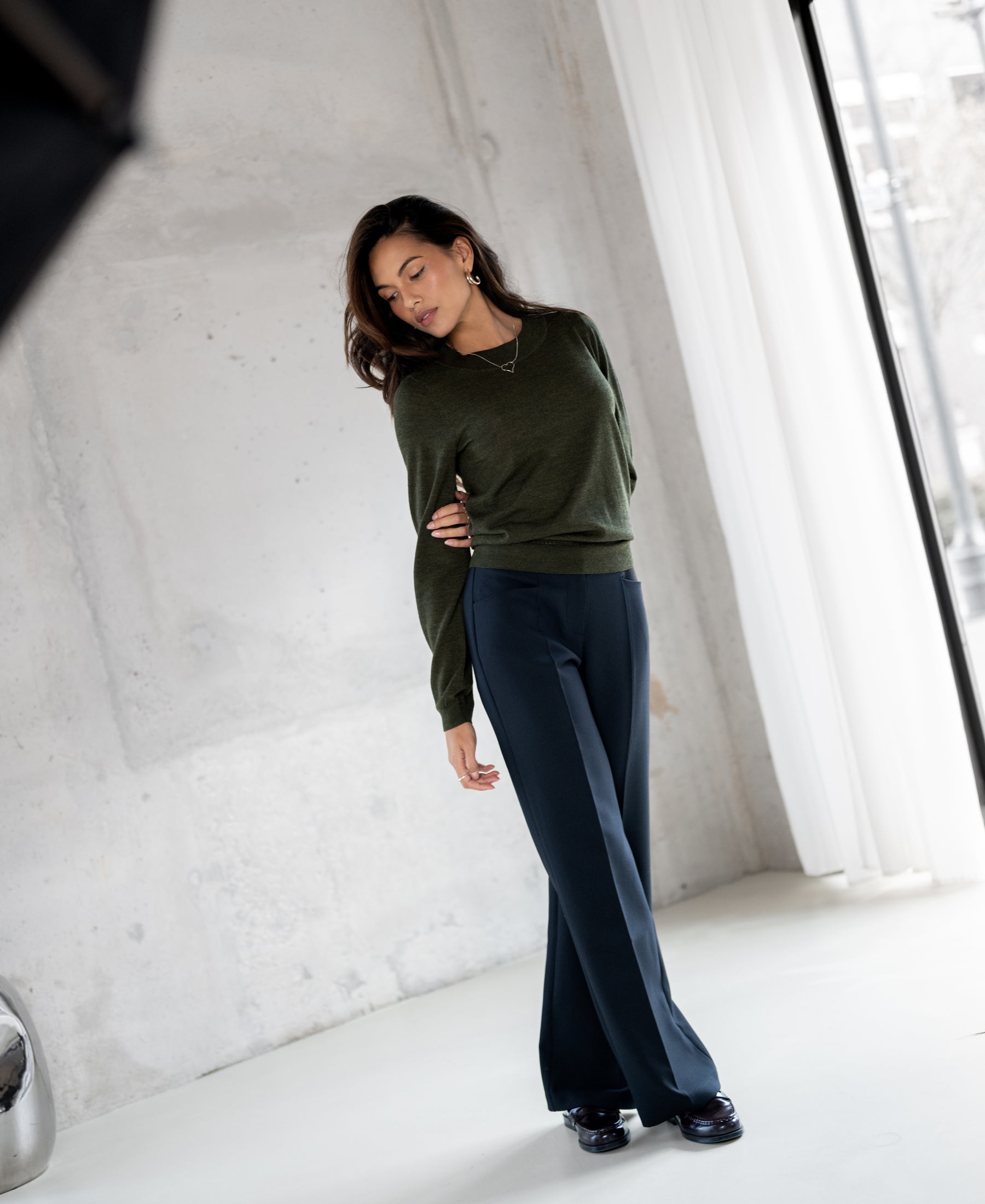 Woman wears the PLEIN PUBLIQUE LA GIGI Merino wool sweater in Bottlegreen, lit by natural window light.