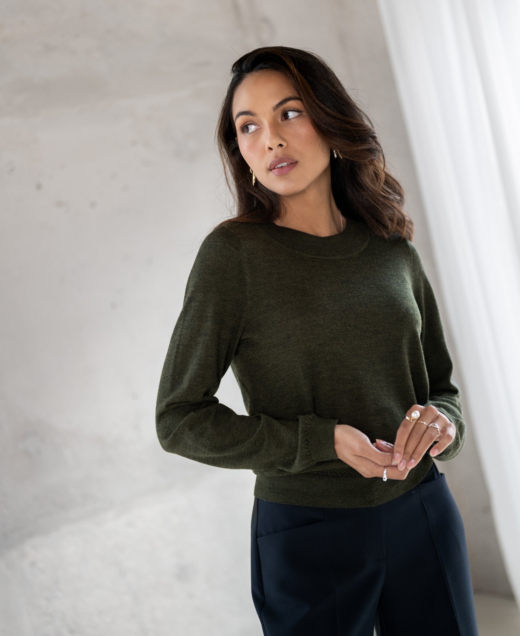 Woman wearing a PLEIN PUBLIQUE merino wool sweater in bottle green, standing near a light curtain.
