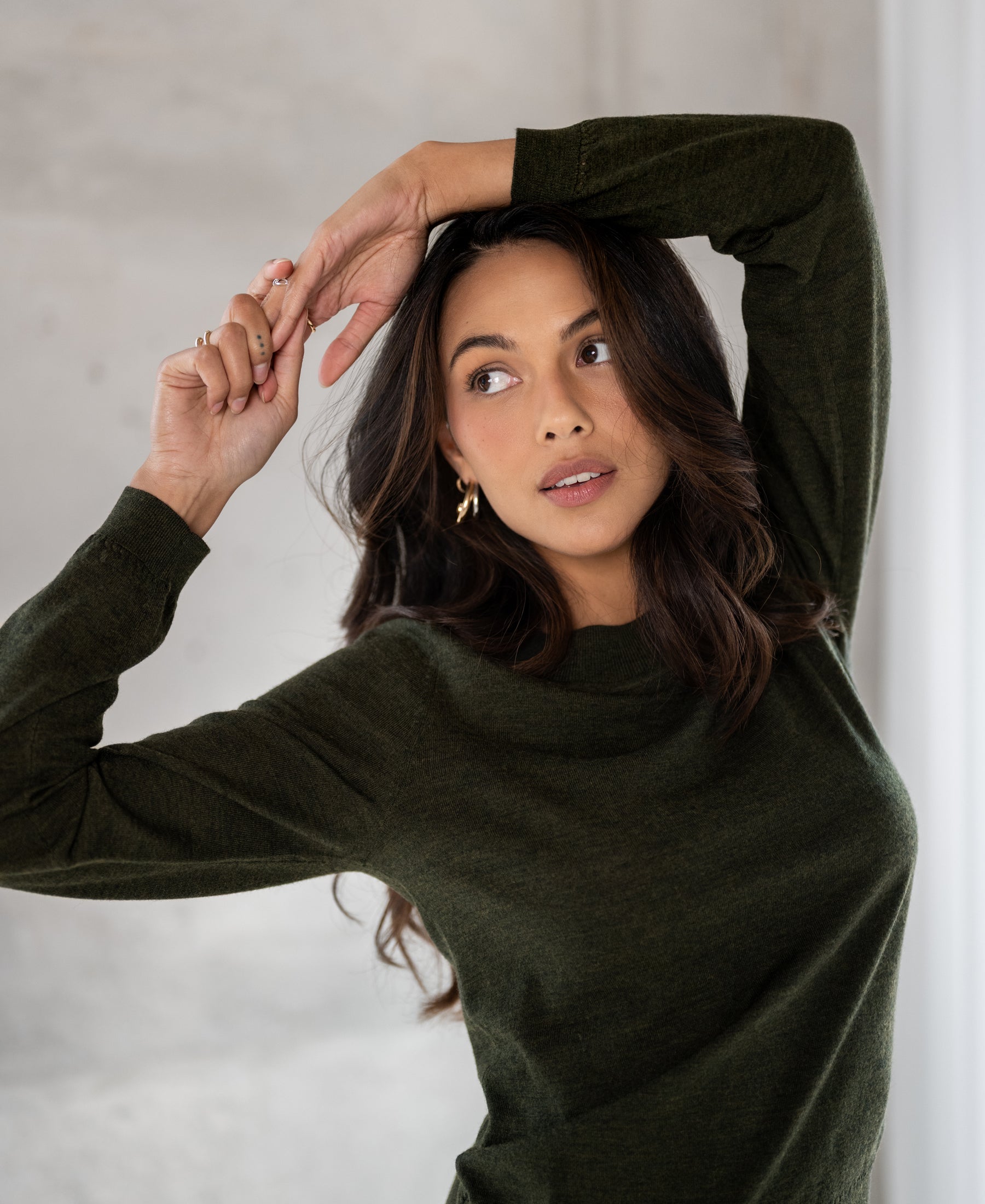 Merino wool sweater in bottle green, shown indoors with arms raised and looking aside.