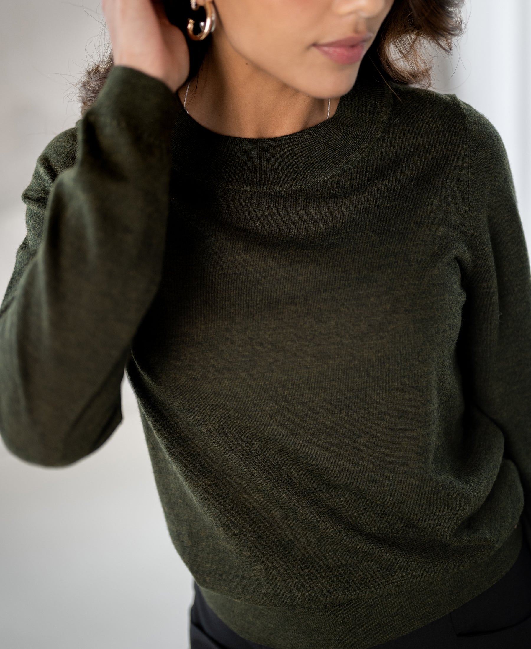 Woman wears the PLEIN PUBLIQUE merino wool sweater LA GIGI in bottle green, blurred background.