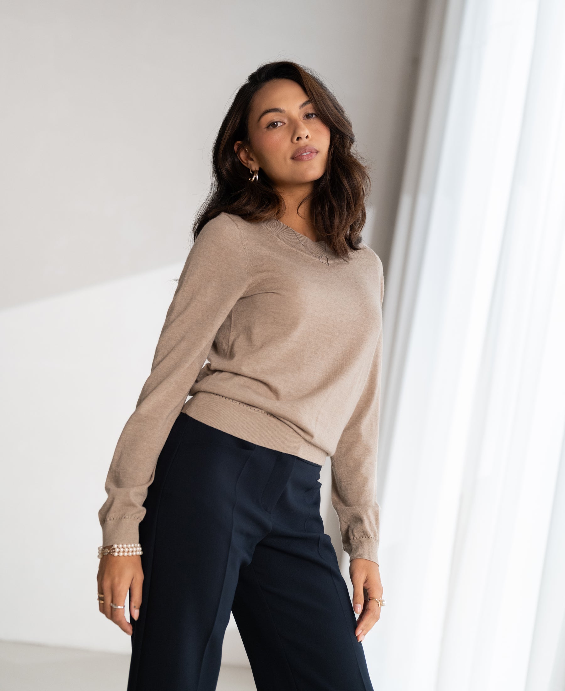 Woman wearing a PLEIN PUBLIQUE Merino wool LA GIGI sweater in sand color stands near a white curtain.
