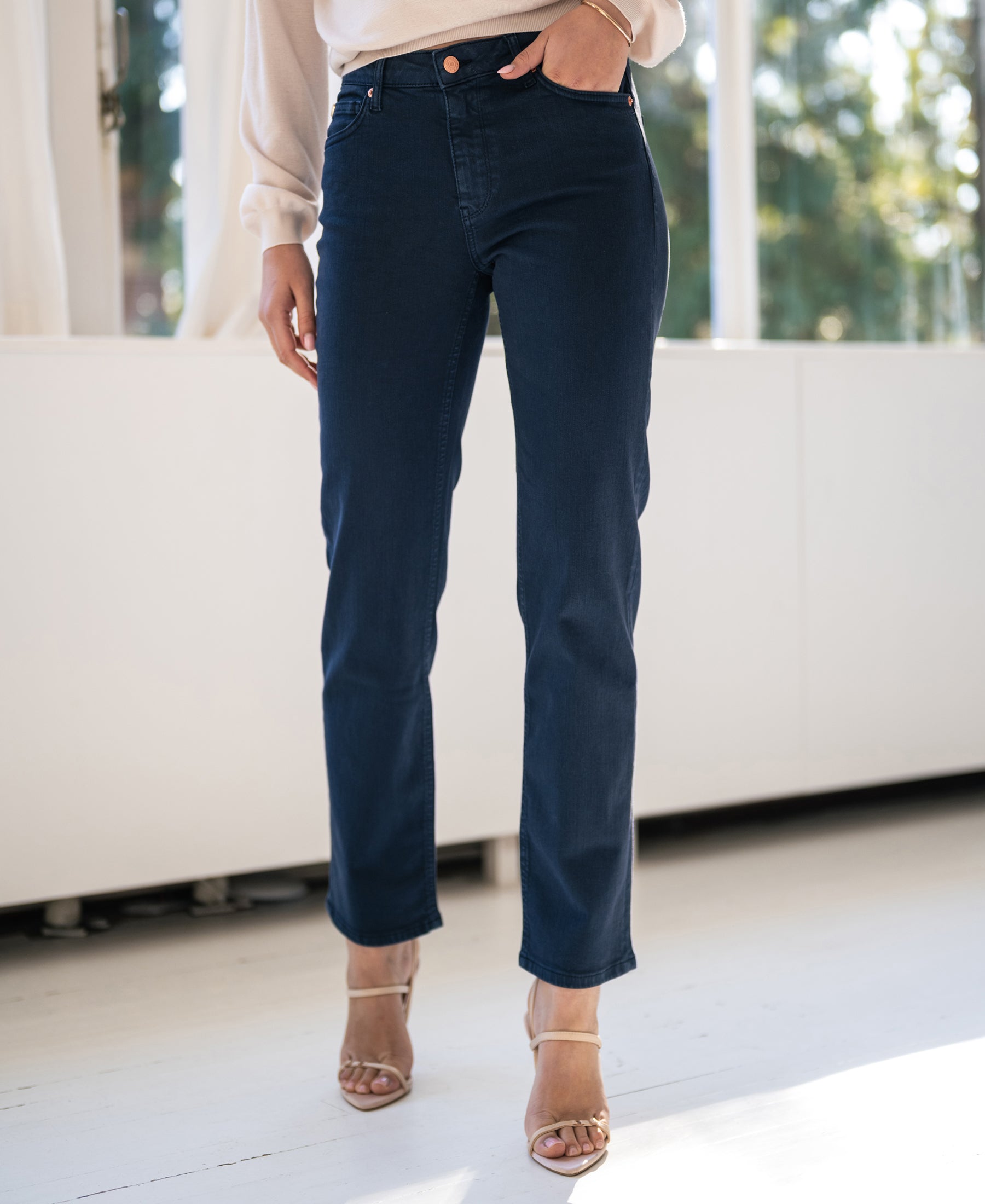 Model wearing dark blue PLEIN PUBLIQUE cropped jeans, standing by a sunlit window indoors.