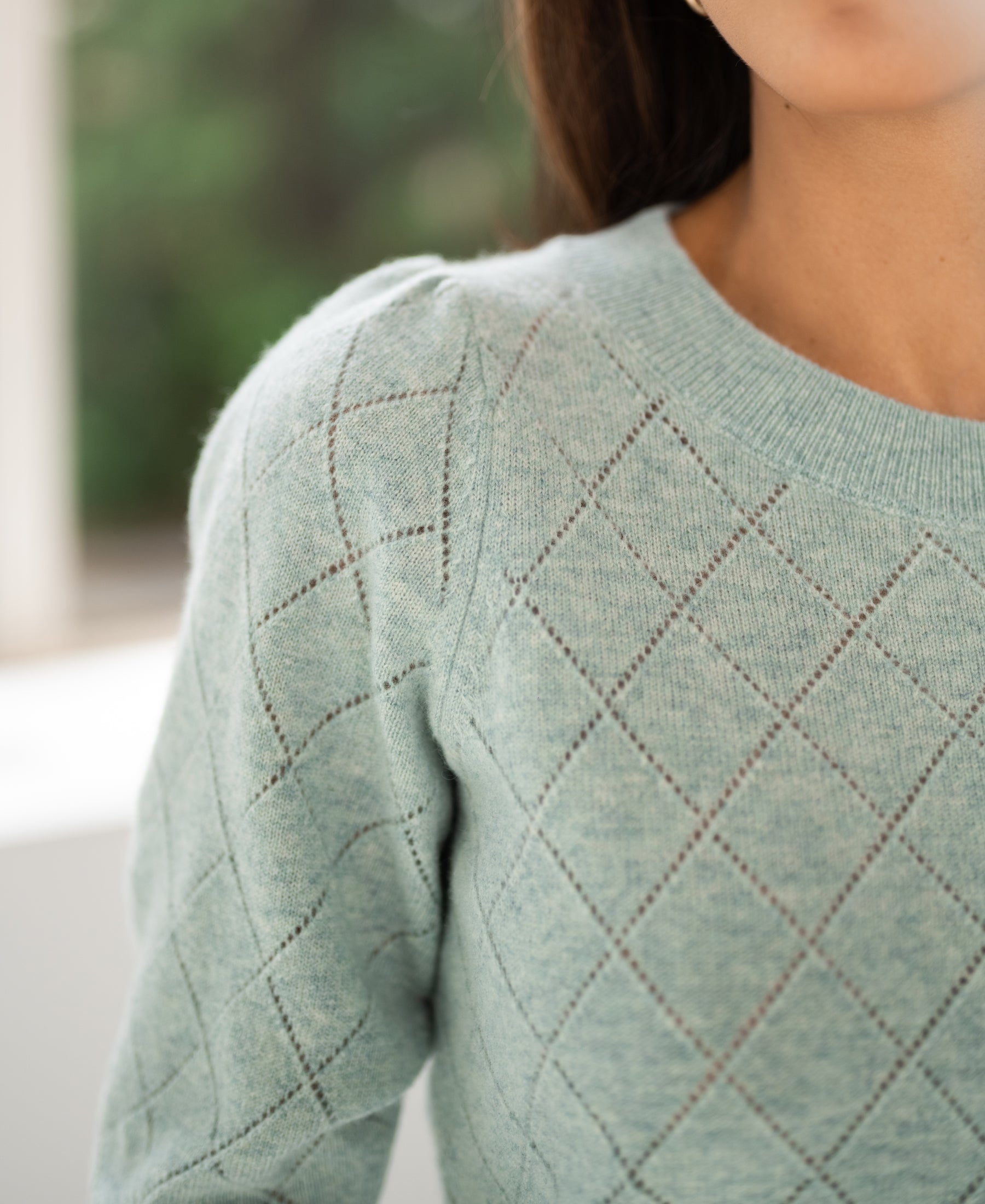 Close-up of an argyle merino wool LE HARVARD sweater in mist green, with the wearers face partially out of frame.