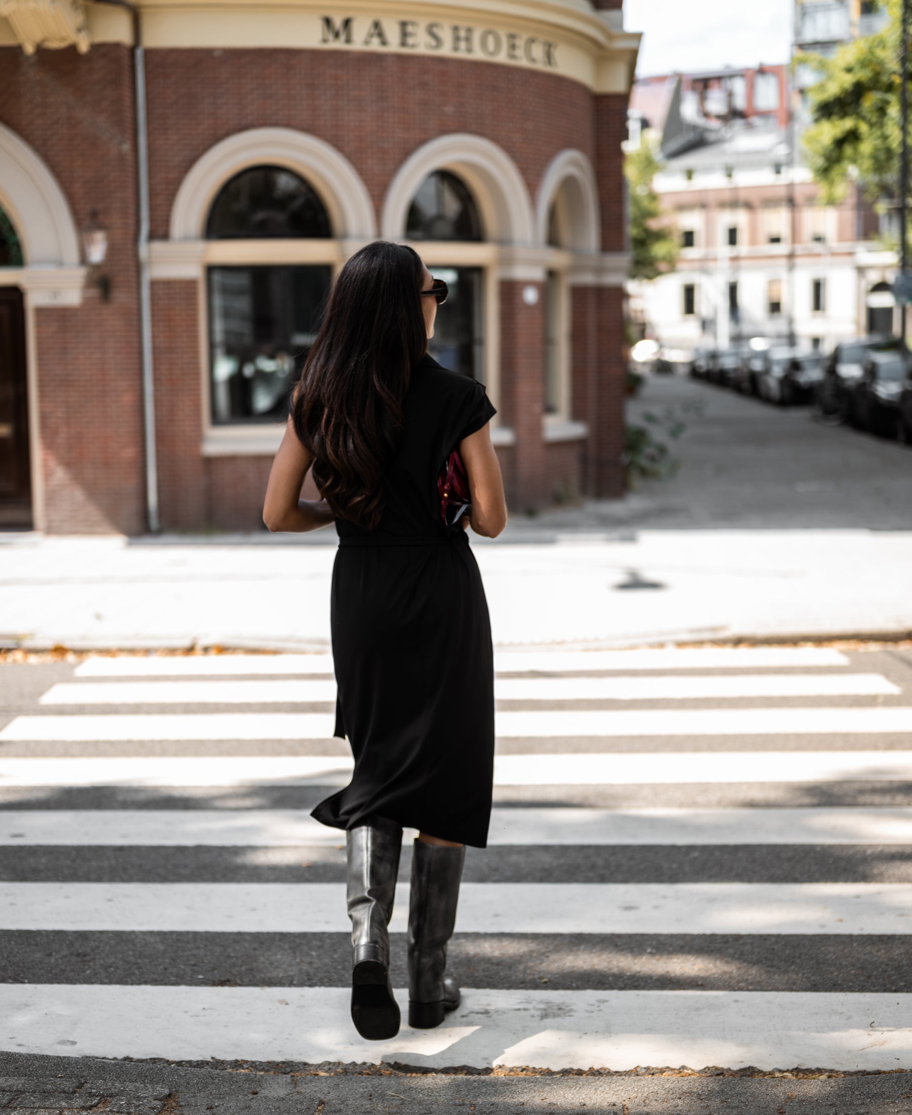 Woman wearing a black INES travel polo dress by PLEIN PUBLIQUE crossing a street by a brick building.
