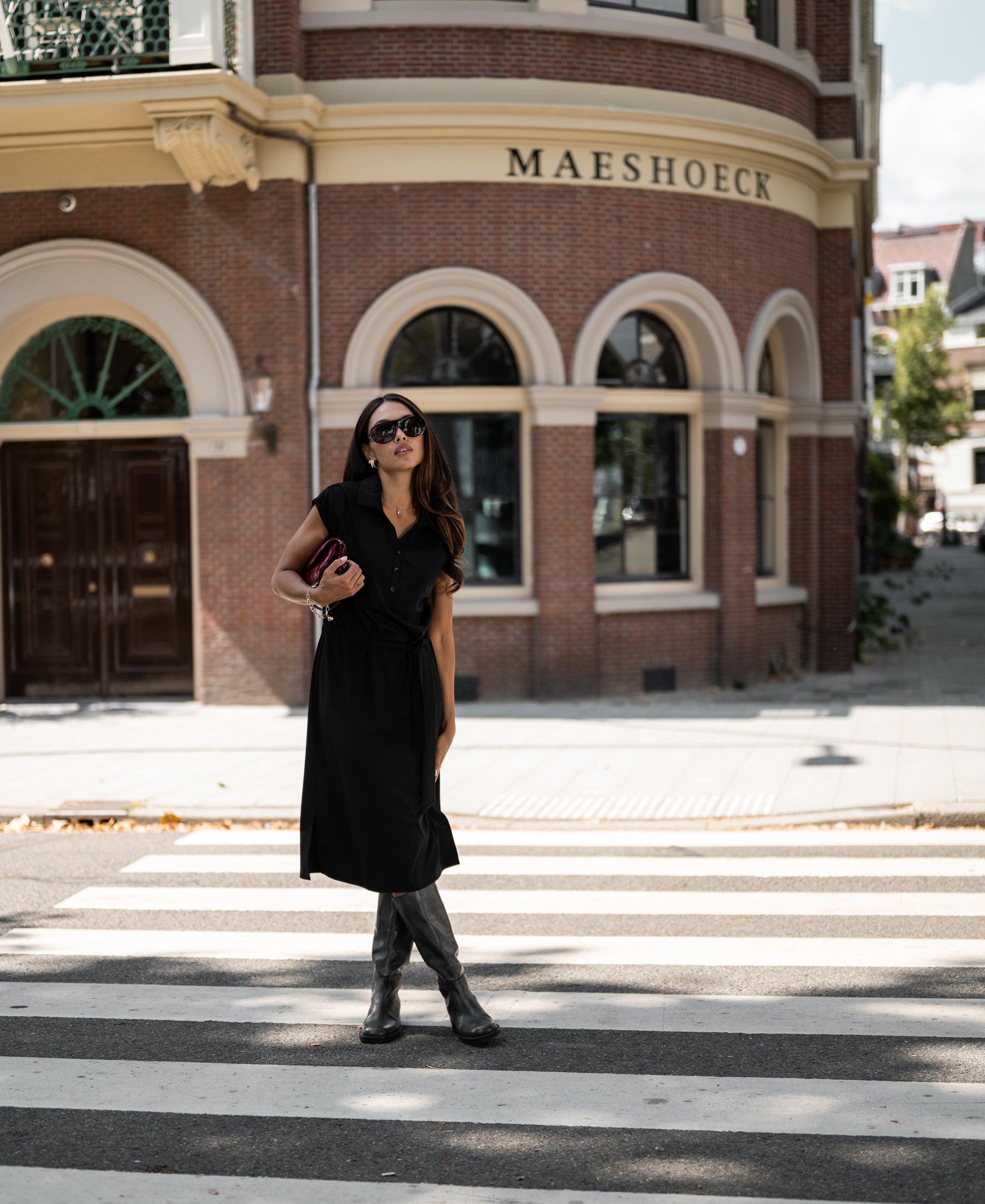 Woman wearing a black PLEIN PUBLIQUE INES travel polo dress stands outside a brick building.